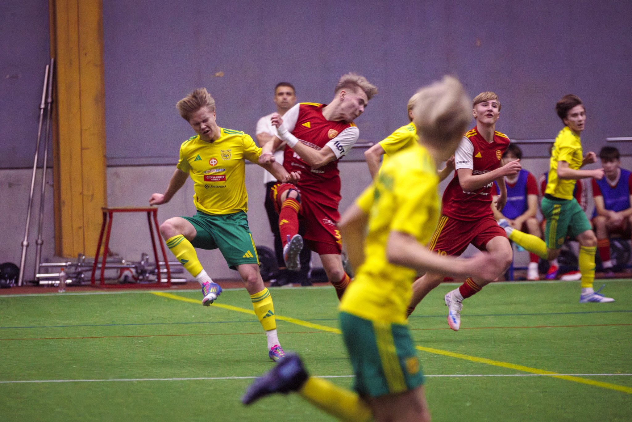 Young boys playing indoor soccer, some in yellow and green uniforms, others in red and yellow uniforms, on a green turf field with a plain background.