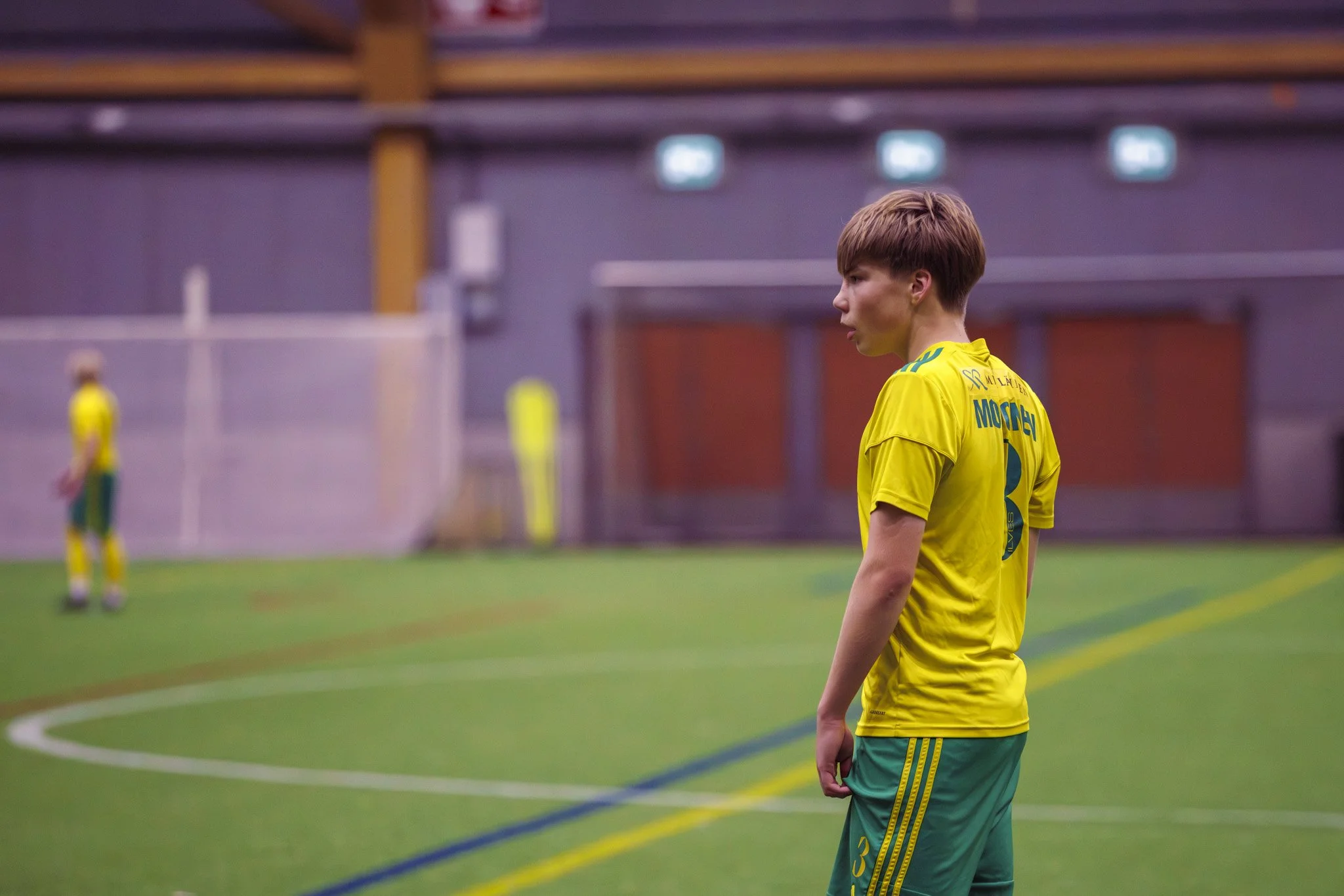 A young male soccer player wearing a yellow and green uniform standing on an indoor soccer field, focused and looking to the right, with another player blurred in the background.