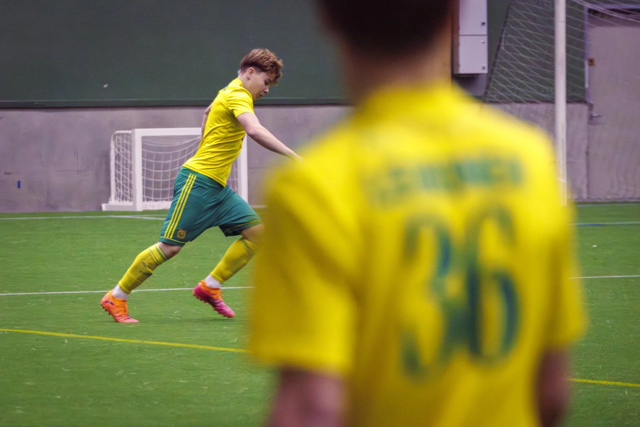 Two young soccer players in yellow and green uniforms practice indoor soccer on a green turf field. One player is in the background near the goal, while the other, in the foreground, is out of focus and facing away from the camera.