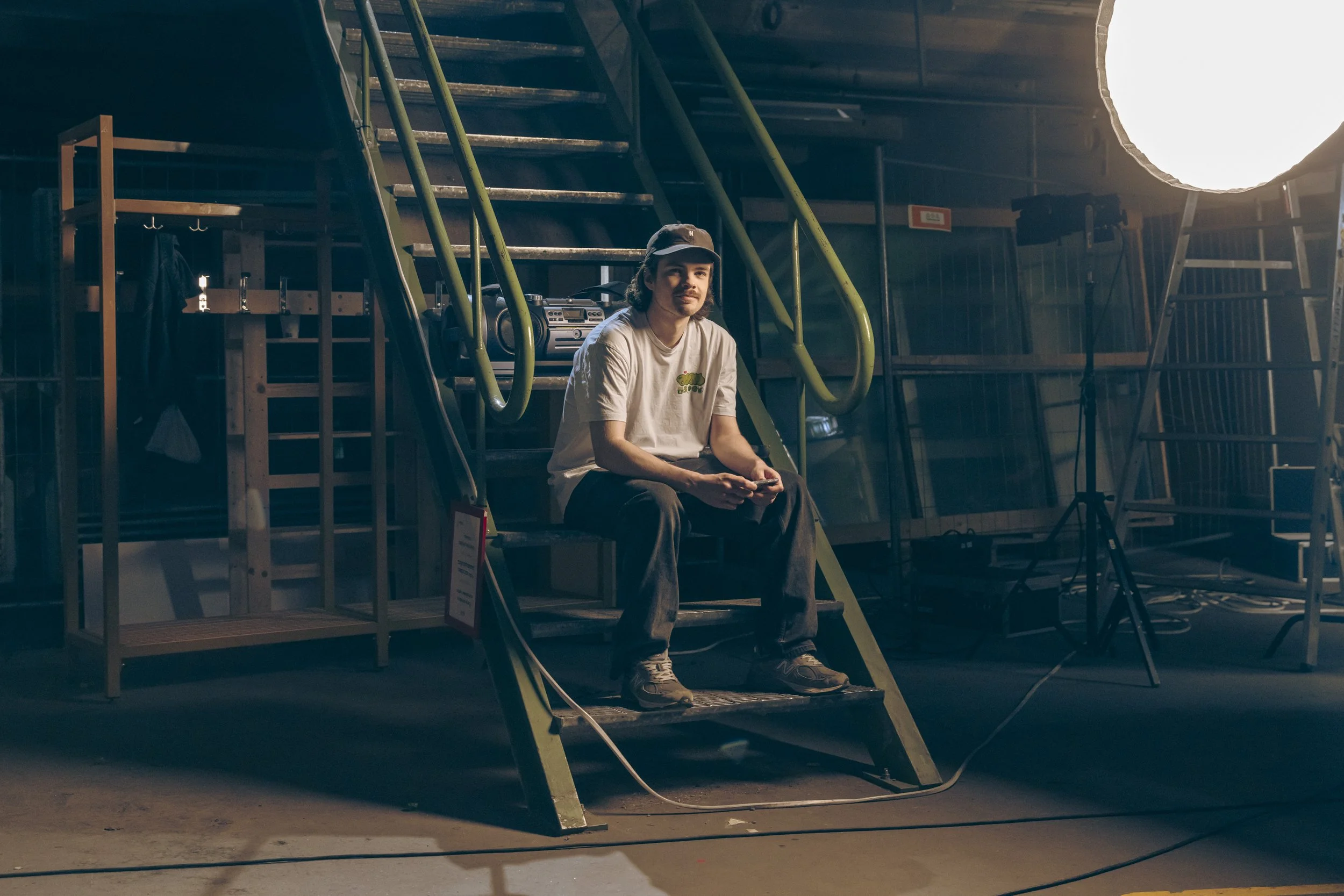 A young man sitting on a staircase in a studio, holding a camera or phone, with film equipment and lighting around him.