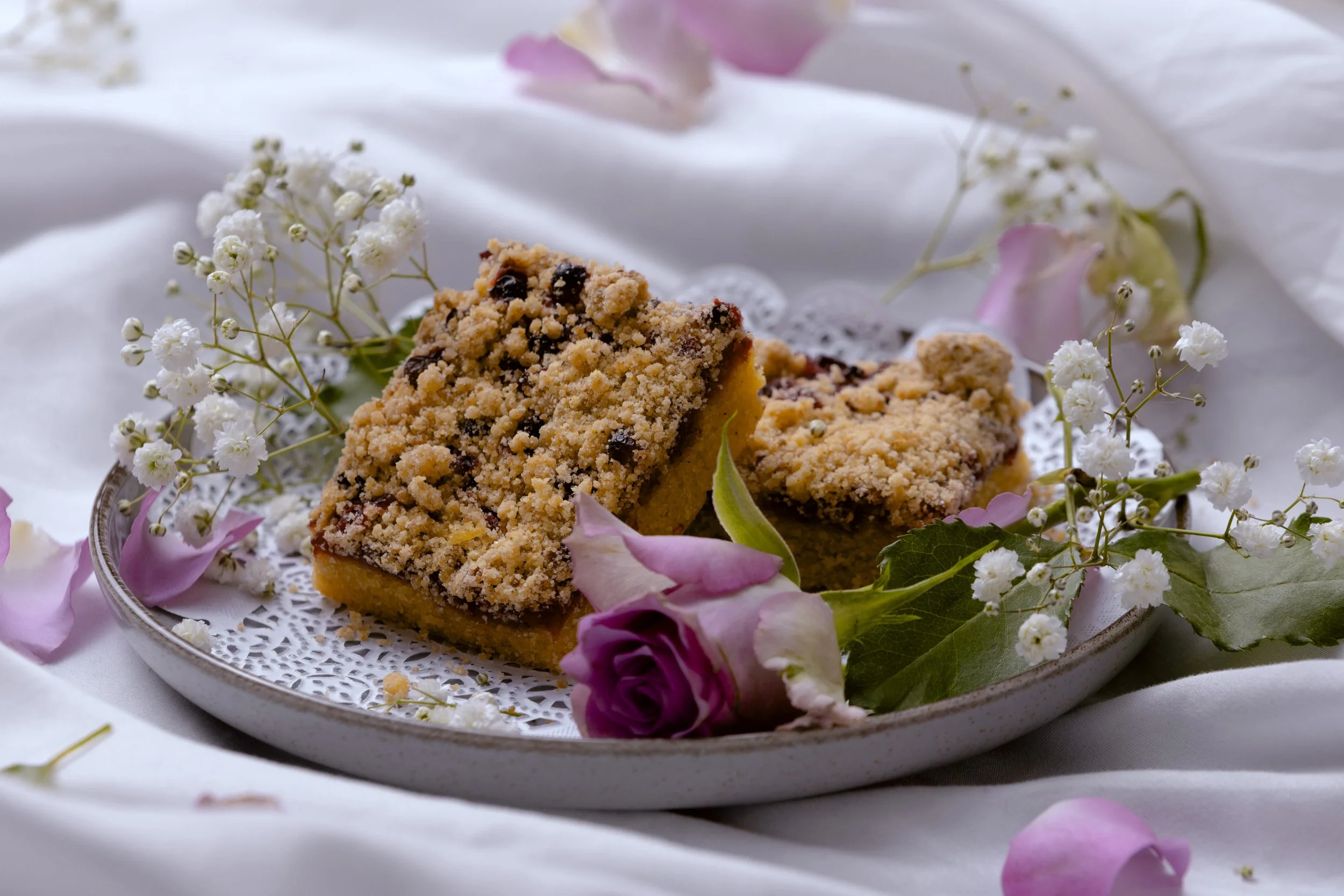 Two crumb-topped fruit bars on a decorative plate, surrounded by pink roses and white baby's breath flowers, on a white cloth background.