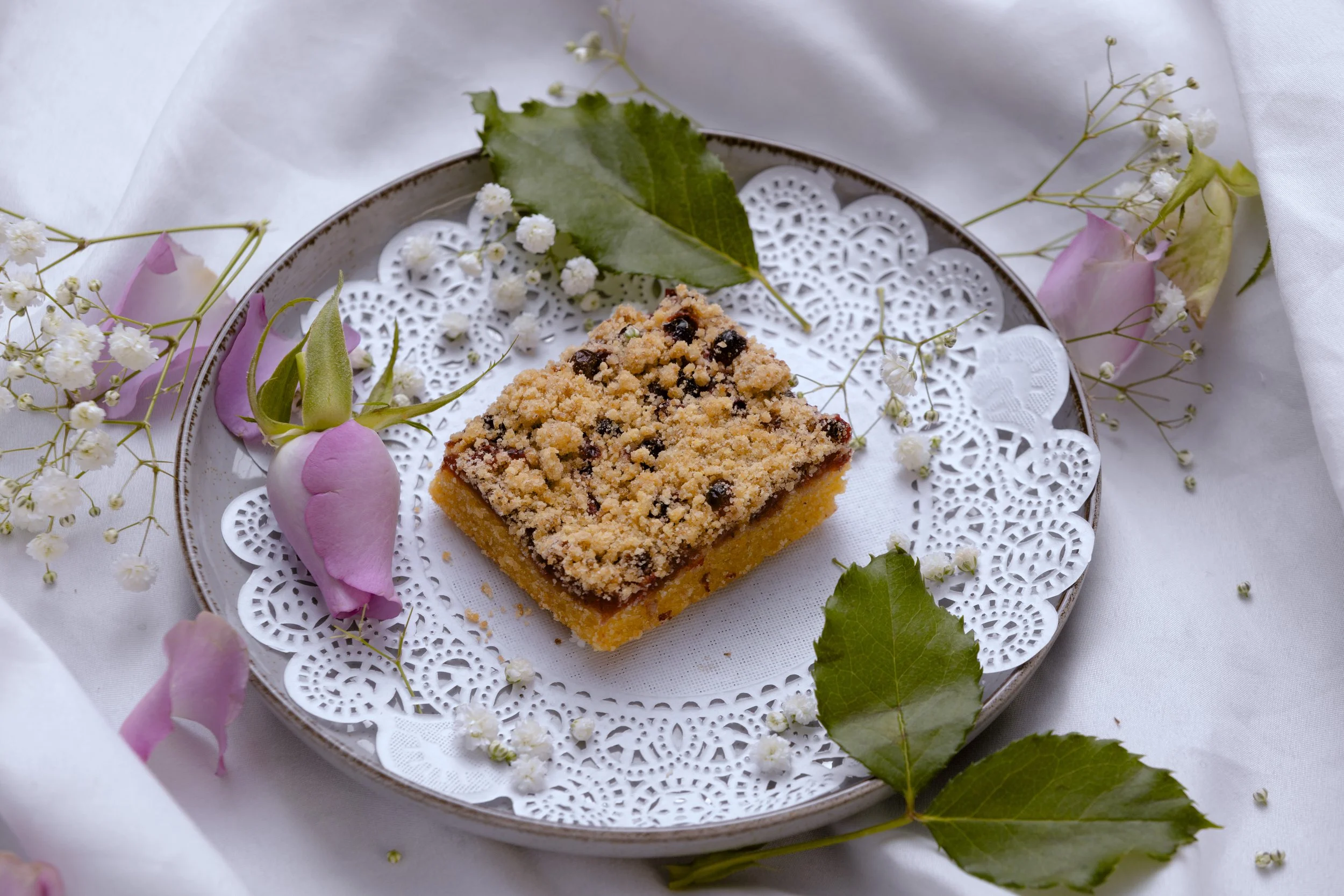 A square piece of crumb-topped fruit crumble on a decorative plate surrounded by pink roses, white baby's breath flowers, and green leaves, on a white tablecloth.