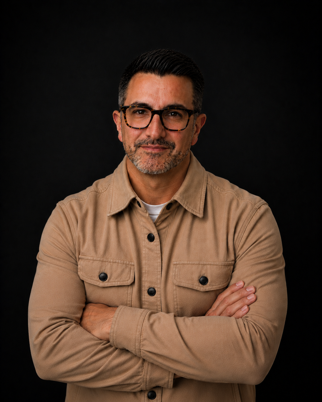 A middle-aged man with dark hair, glasses, and a closely trimmed beard, wearing a beige jacket, standing against a black background with arms crossed.