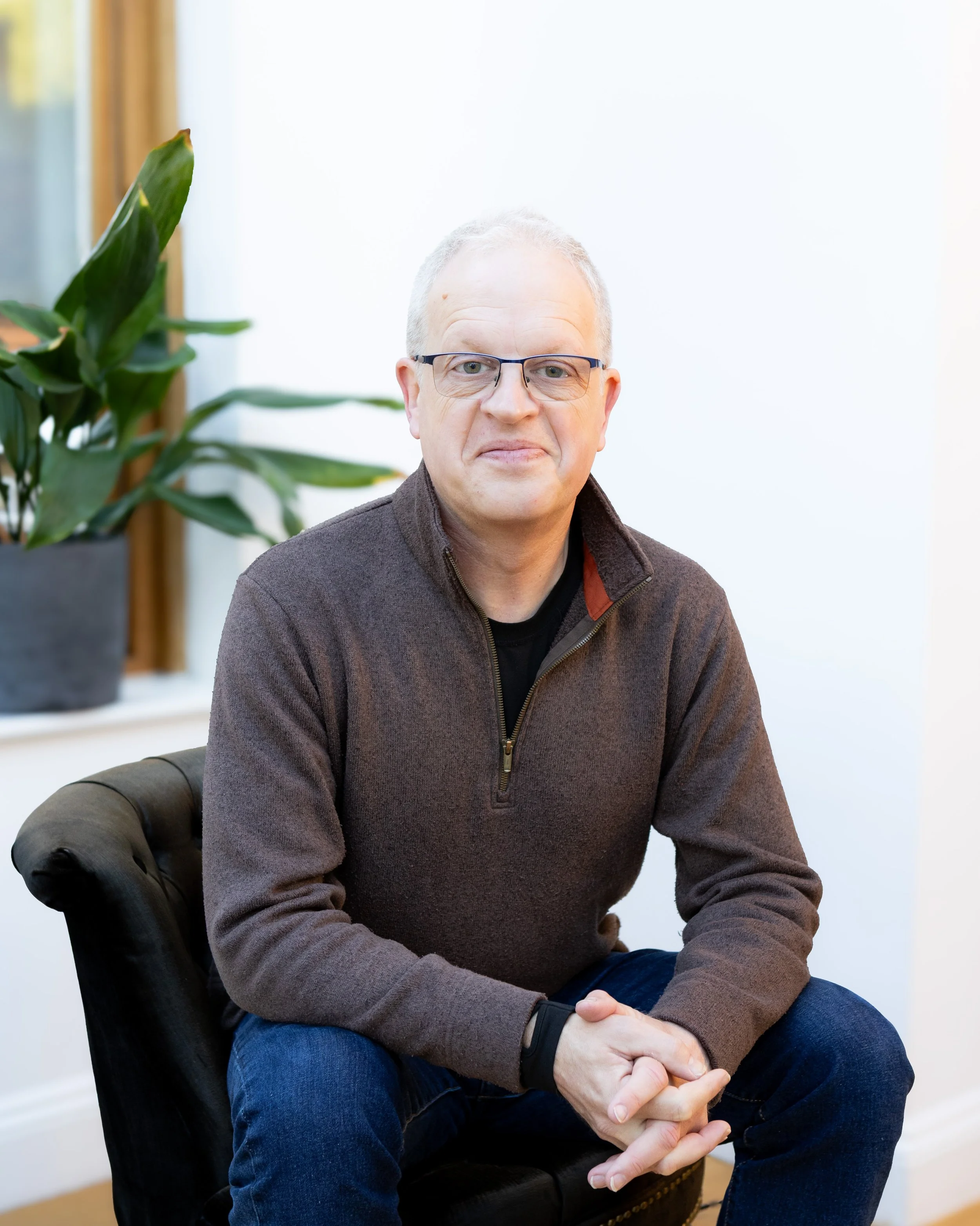 A middle-aged man with glasses and short, light-colored hair sitting on a dark leather chair indoors, wearing a brown quarter-zip sweater and blue jeans, with a large green plant in the background.