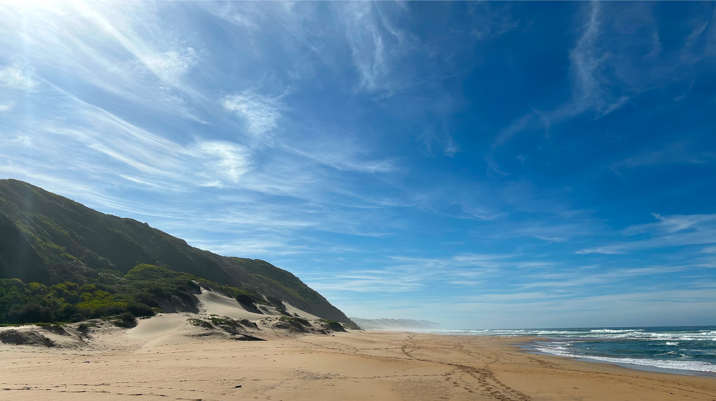 Sandy beach with ocean waves and grassy dunes under a blue sky with clouds.