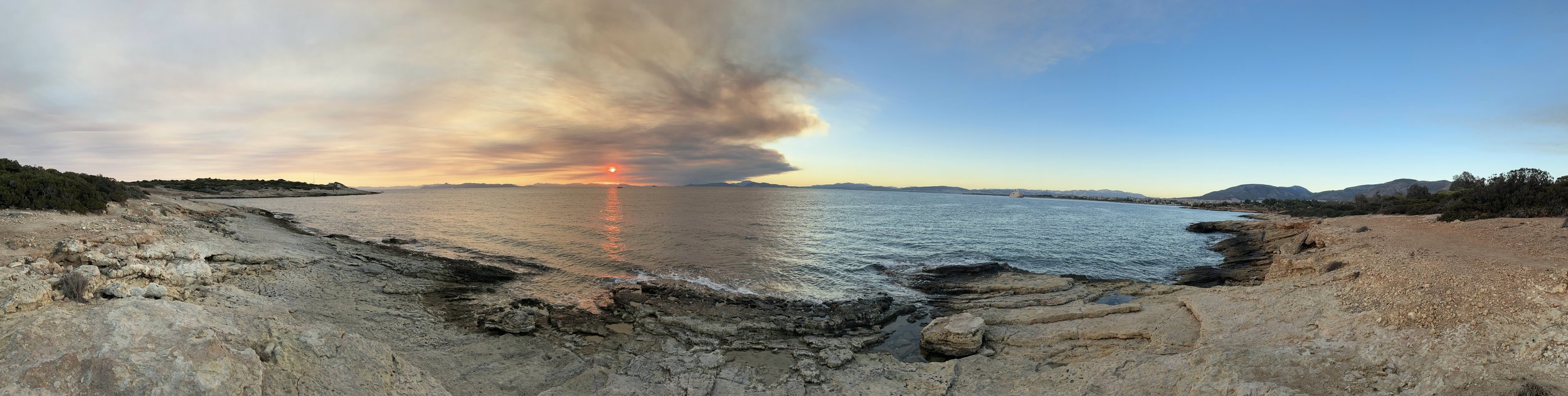 Coastal landscape with rocky shoreline, calm sea, and sunset creating a reflection on the water, surrounded by distant hills and clear skies. Taken at Pikpa Beach in Athens, Greece.