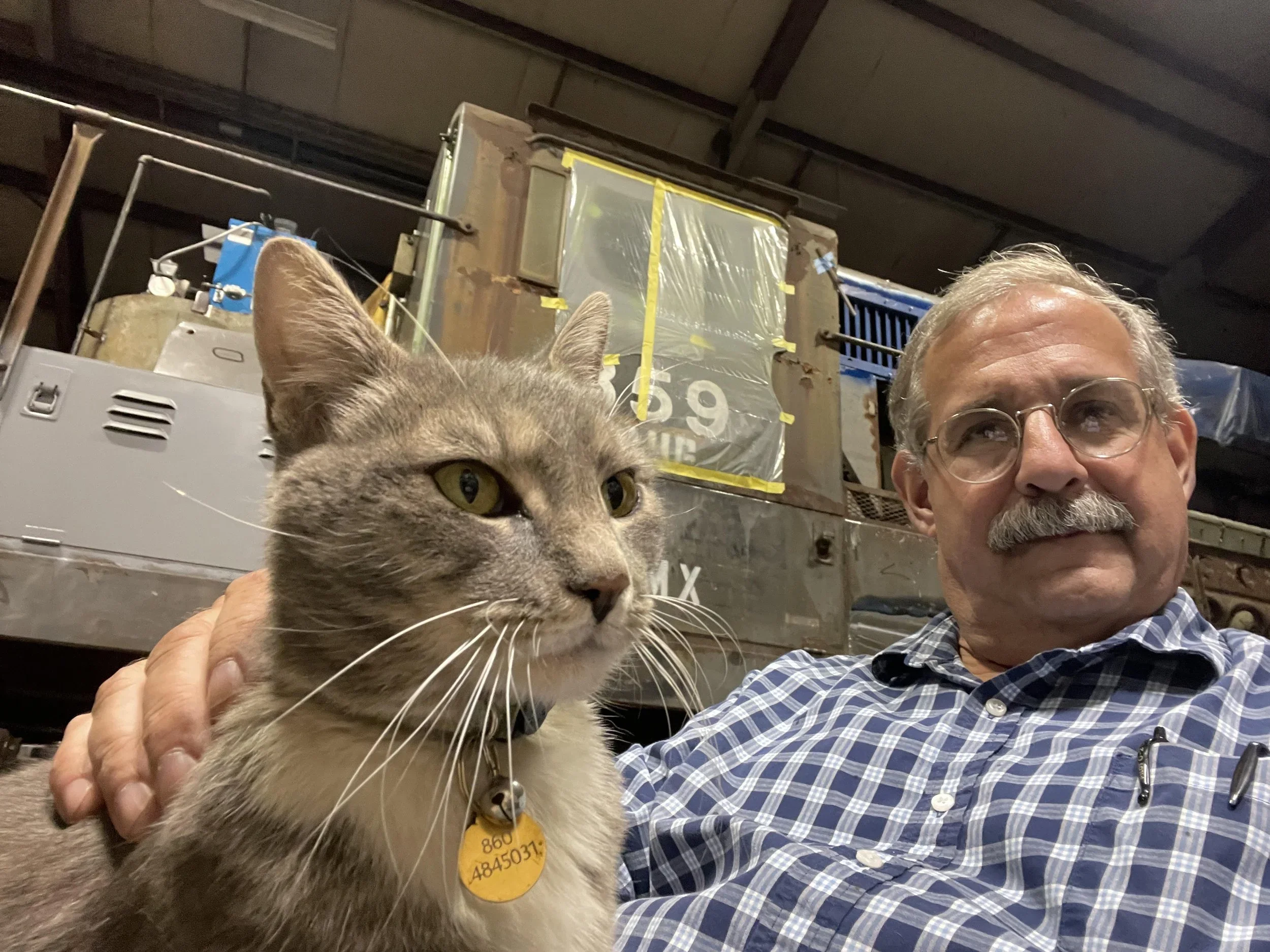 A man with glasses and a mustache wearing a plaid shirt taking a selfie with a gray tabby cat wearing a yellow ID collar, in an industrial setting with metal equipment and machinery in the background.