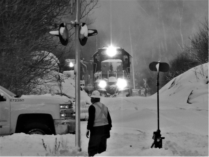 Black and white photo of a railroad worker by a signal in the snow with an engine