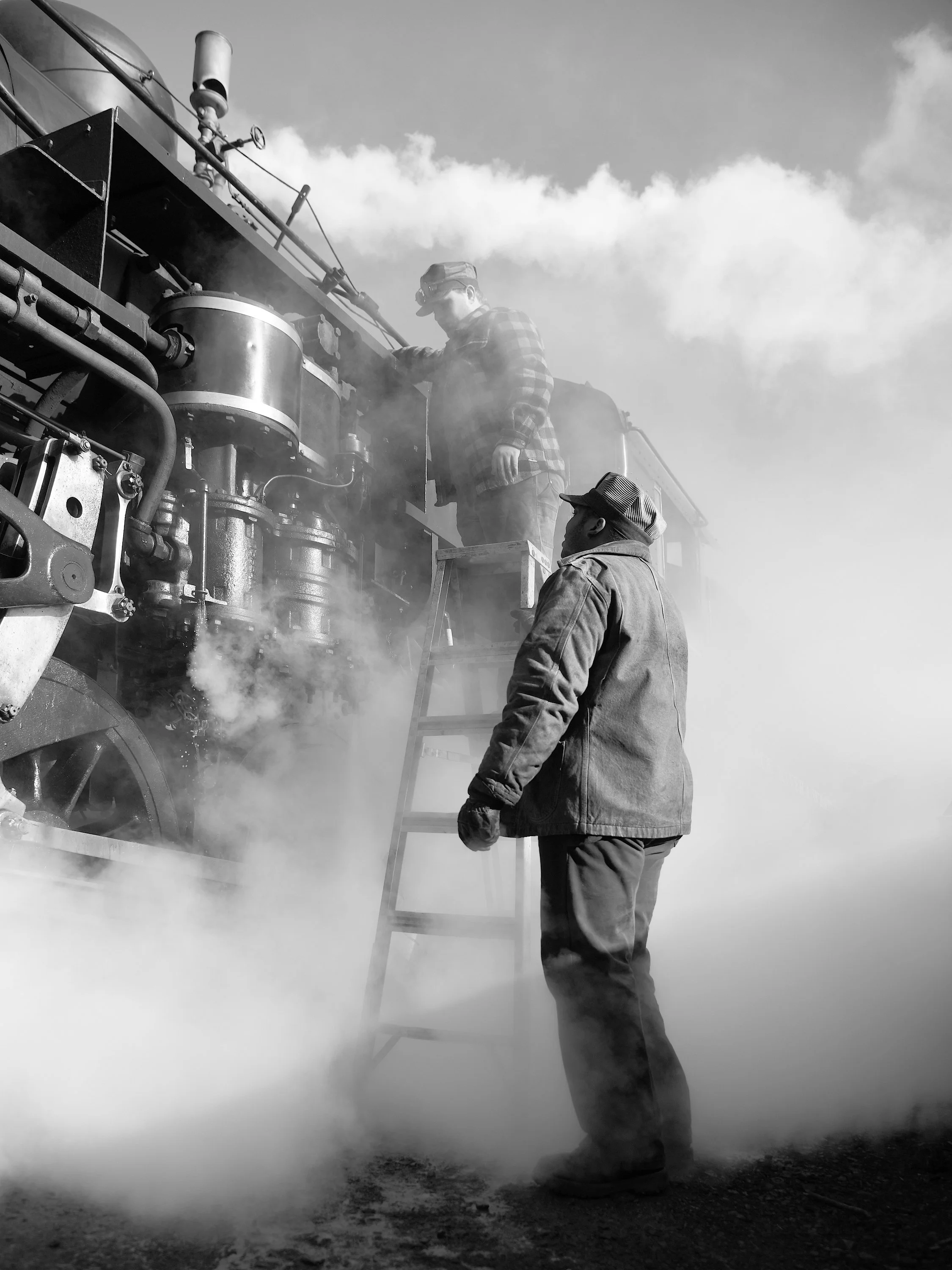 Two men working on a steam train, with one on a ladder inspecting the engine and the other standing beside, surrounded by steam and smoke, outdoors under a cloudy sky.