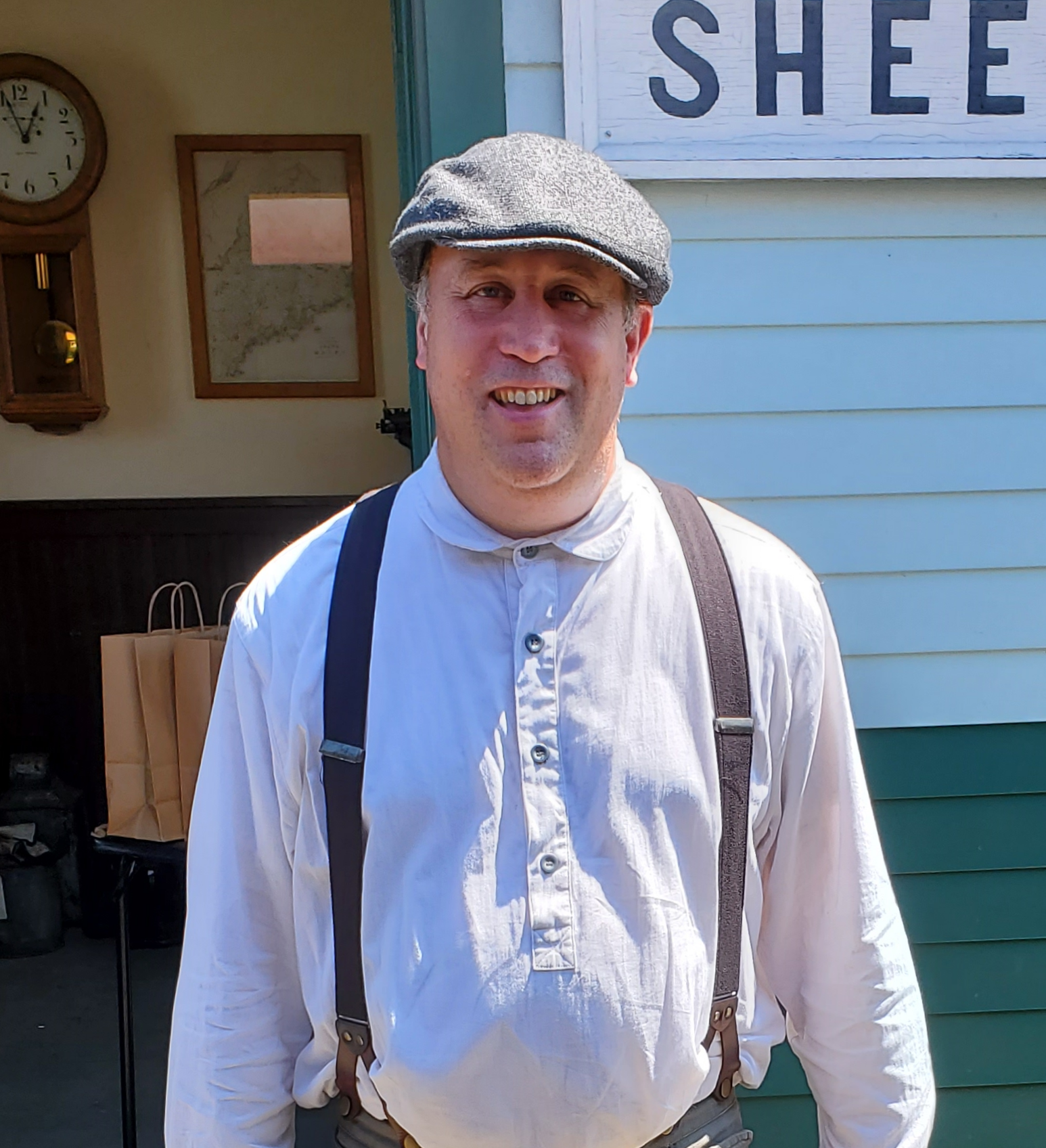 Man wearing a flat cap, white button-up shirt, and suspenders, smiling outdoors with a sign and a clock in the background.