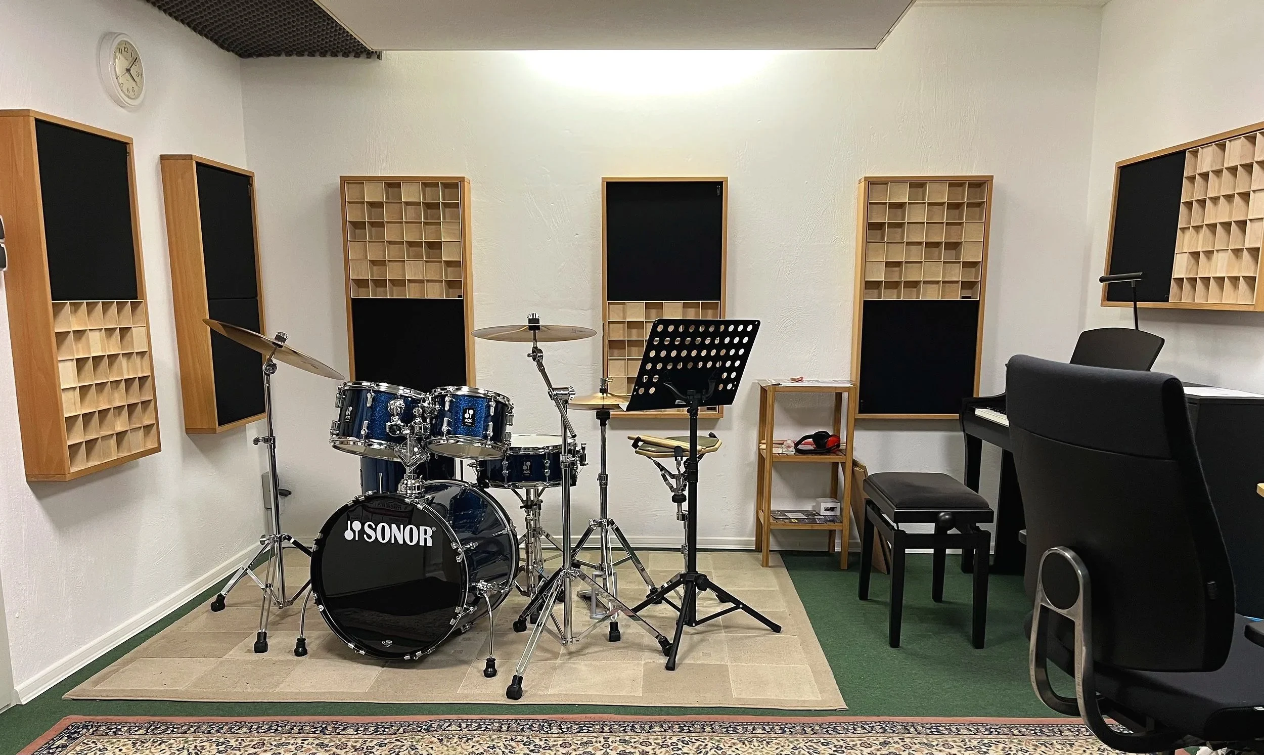 Musikschul Raum mit blauem Schlagzeug auf der linken Seite, schwarzem Klavier auf der rechten Seite und Akustik-Elementen an den Wänden.