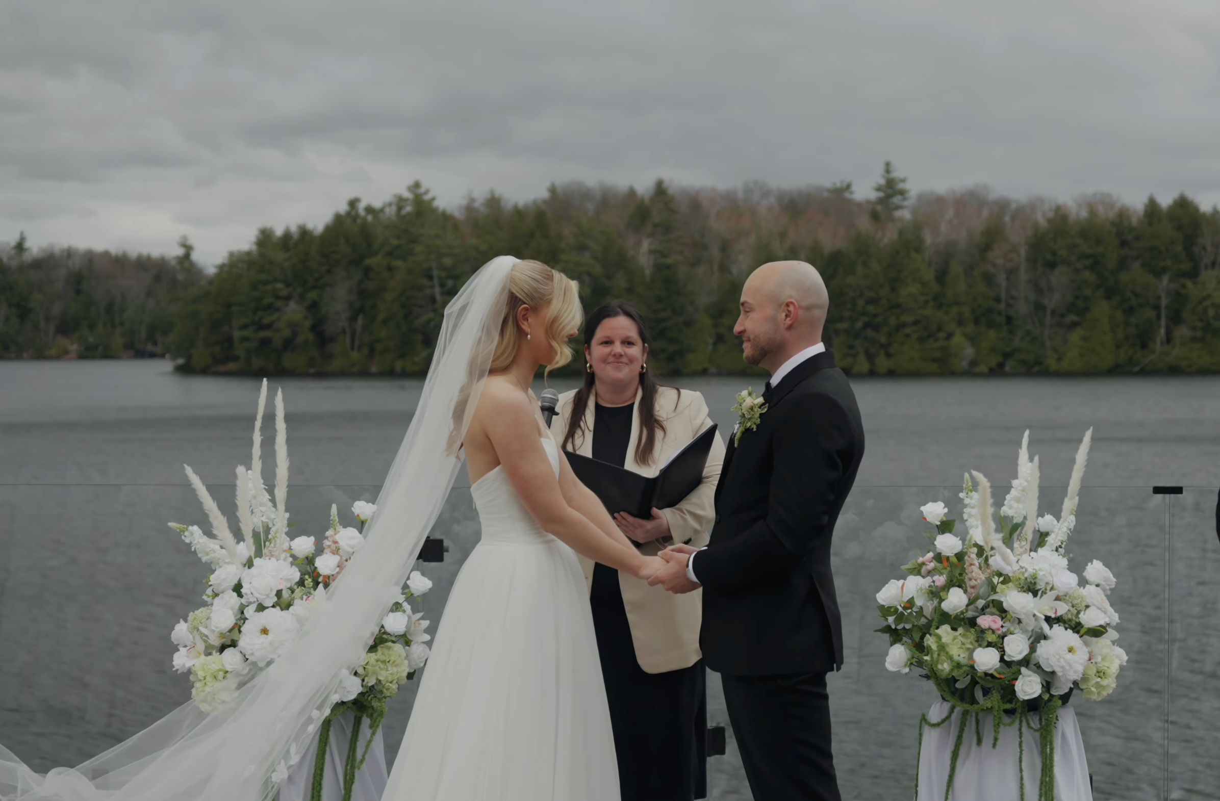 A couple getting married outdoors on a cloudy day by a lake, with a floral altar and officiant present.