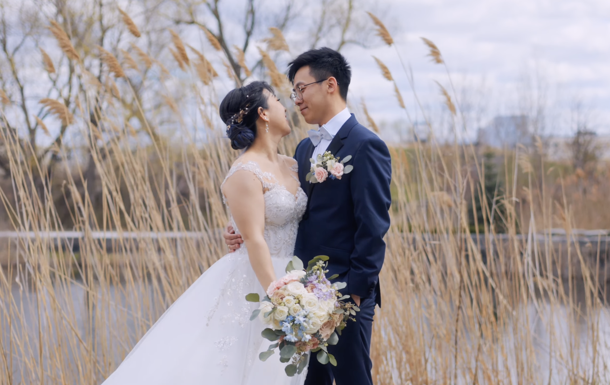A bride and groom holding each other outdoors during their wedding, surrounded by tall dried grass, with trees and a cloudy sky in the background.