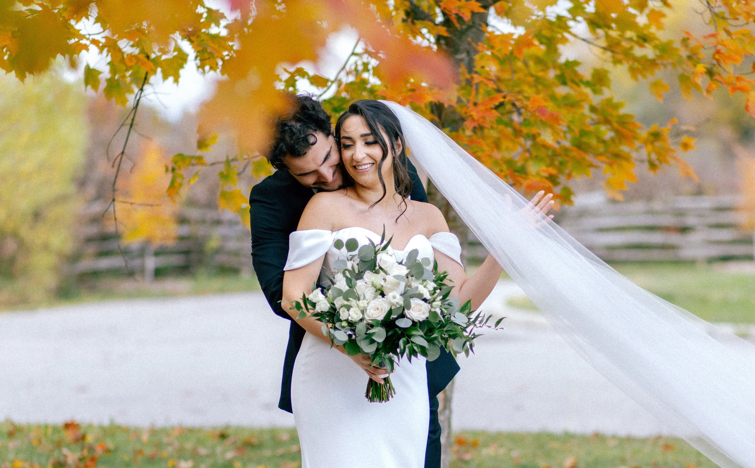 A bride and groom embrace outdoors during fall, with colorful autumn leaves in the background. The bride wears a white off-the-shoulder wedding dress, holding a large bouquet of white and green flowers. The groom is dressed in a dark suit.