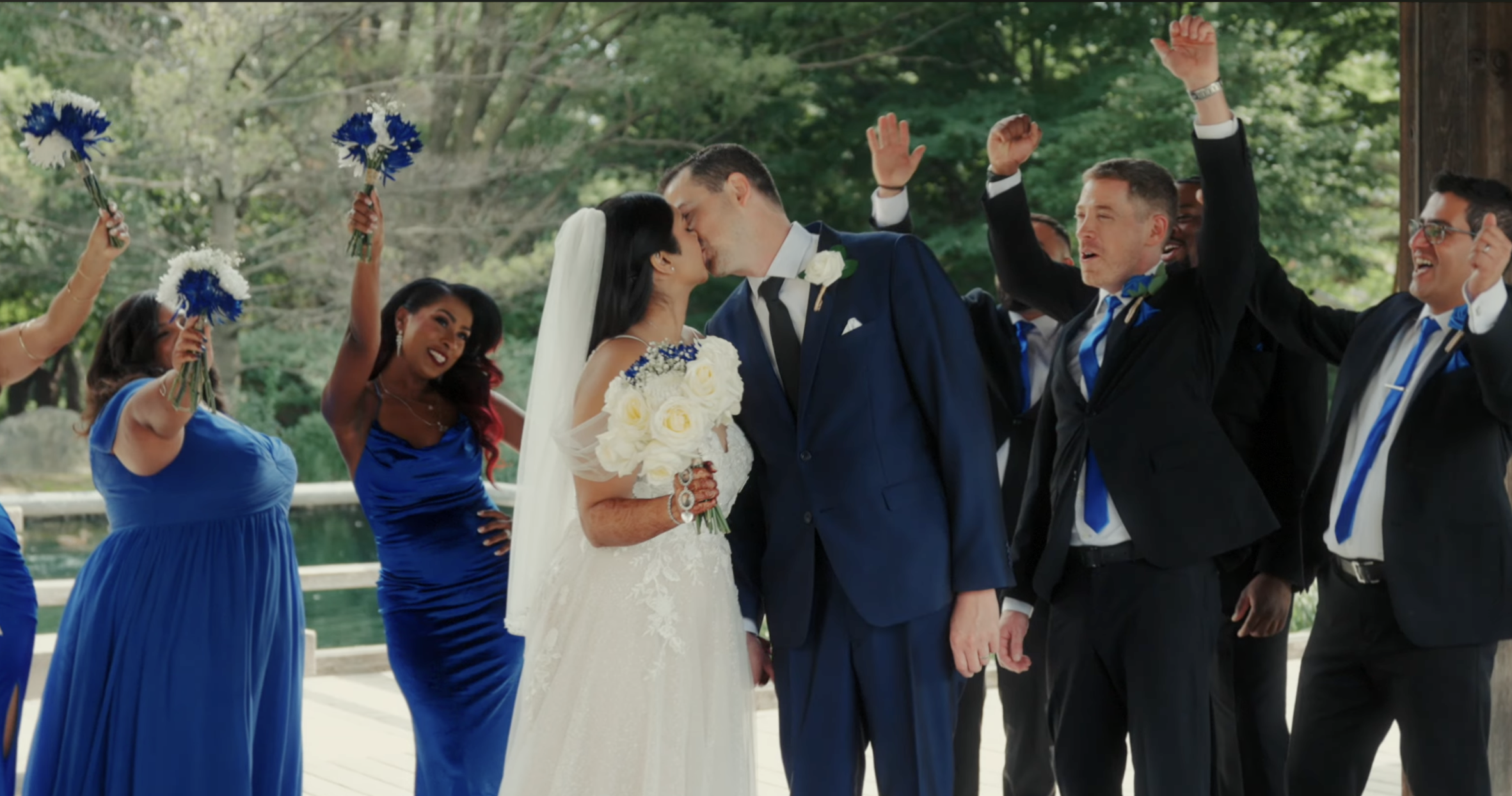 A bride and groom share a kiss during their wedding ceremony, surrounded by bridesmaids and groomsmen cheering, holding bouquets and wearing formal attire outdoors.