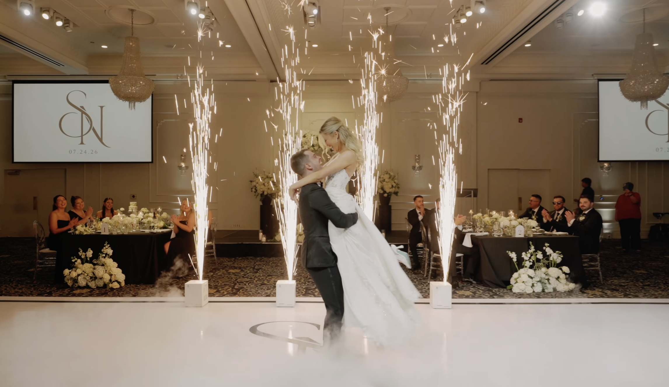 A bride and groom sharing a dance at their wedding reception, surrounded by sparklers and guests clapping, with elegant decor and large screens displaying their initials and wedding date.