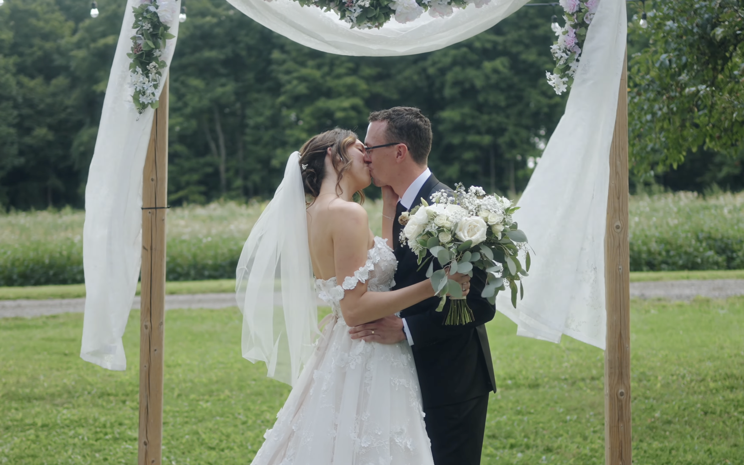 A bride and groom kissing under a wooden wedding arch decorated with white fabric and flowers, in an outdoor setting with green trees in the background.
