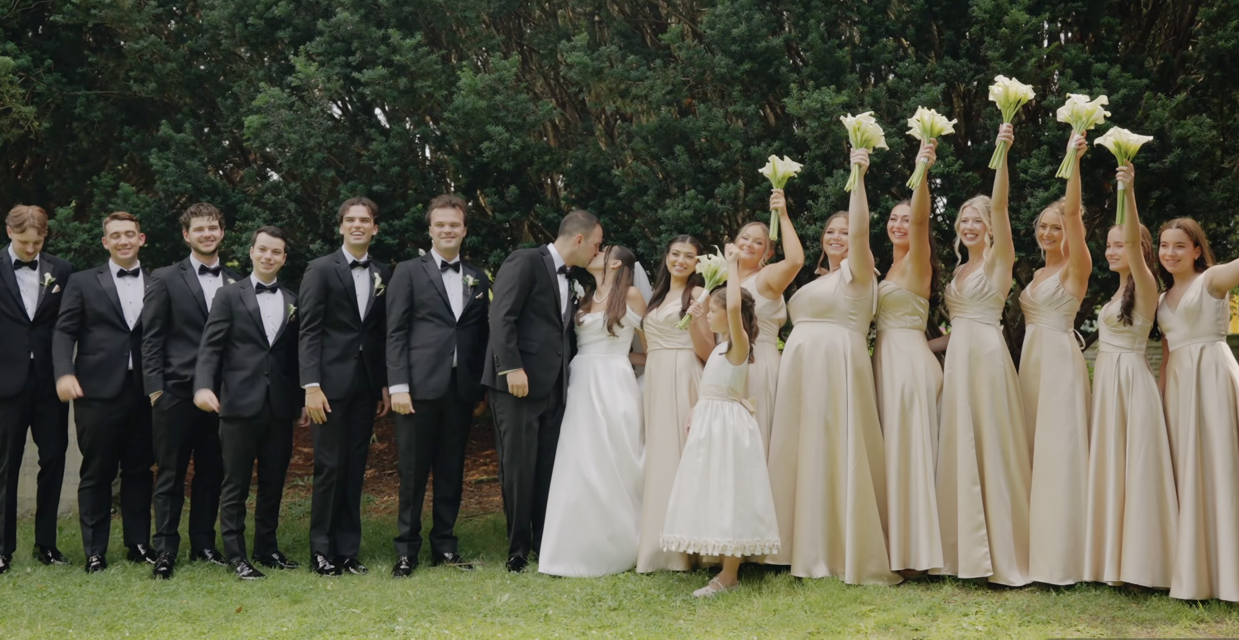 A wedding party standing outdoors in front of trees, with men in black tuxedos and women in cream dresses holding bouquets, some women holding flowers high in the air and a couple in the center sharing a kiss.