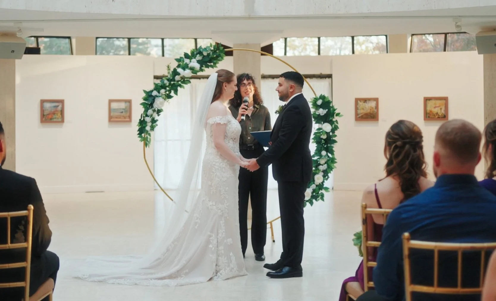 A bride and groom holding hands during their wedding ceremony, standing before an officiant, with guests seated in chairs watching, inside a bright room with artwork on the walls and a decorative floral arch behind them.