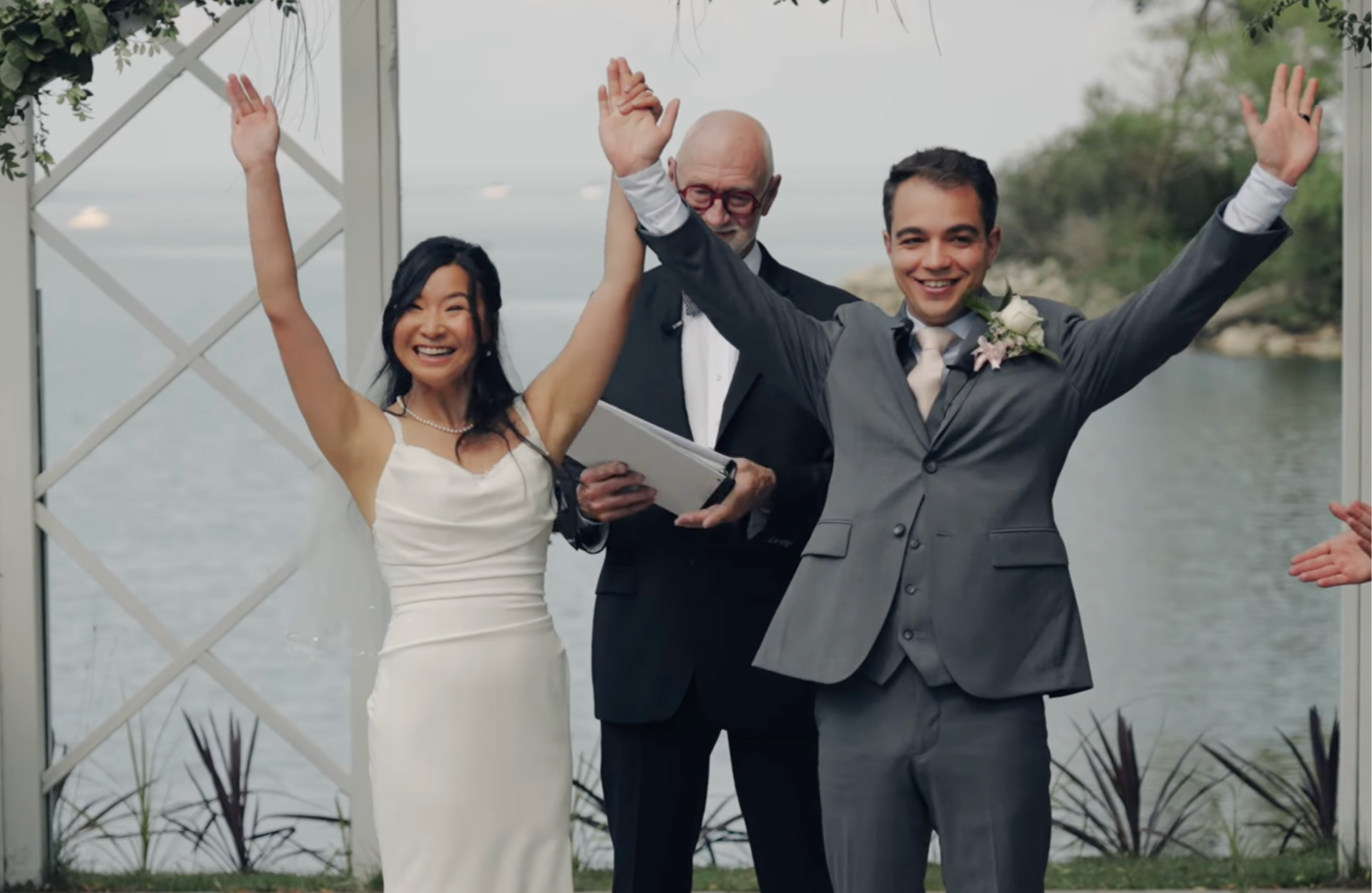 A wedding ceremony outdoors with a bride and groom raising their hands in celebration, accompanied by a man with glasses holding a book, on a lakeside with trees in the background.