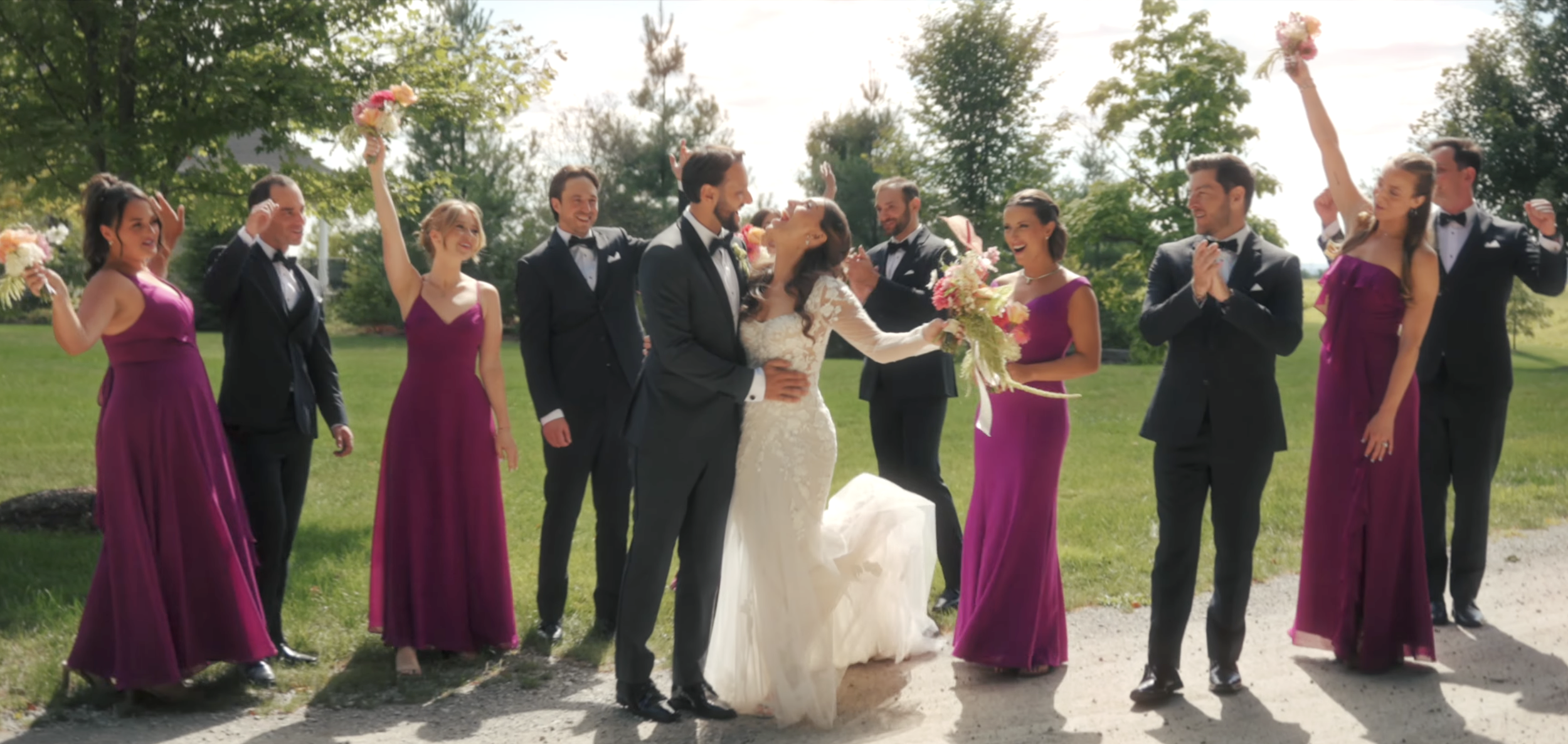 A bride and groom kiss while surrounded by wedding party members outdoors on a sunny day.