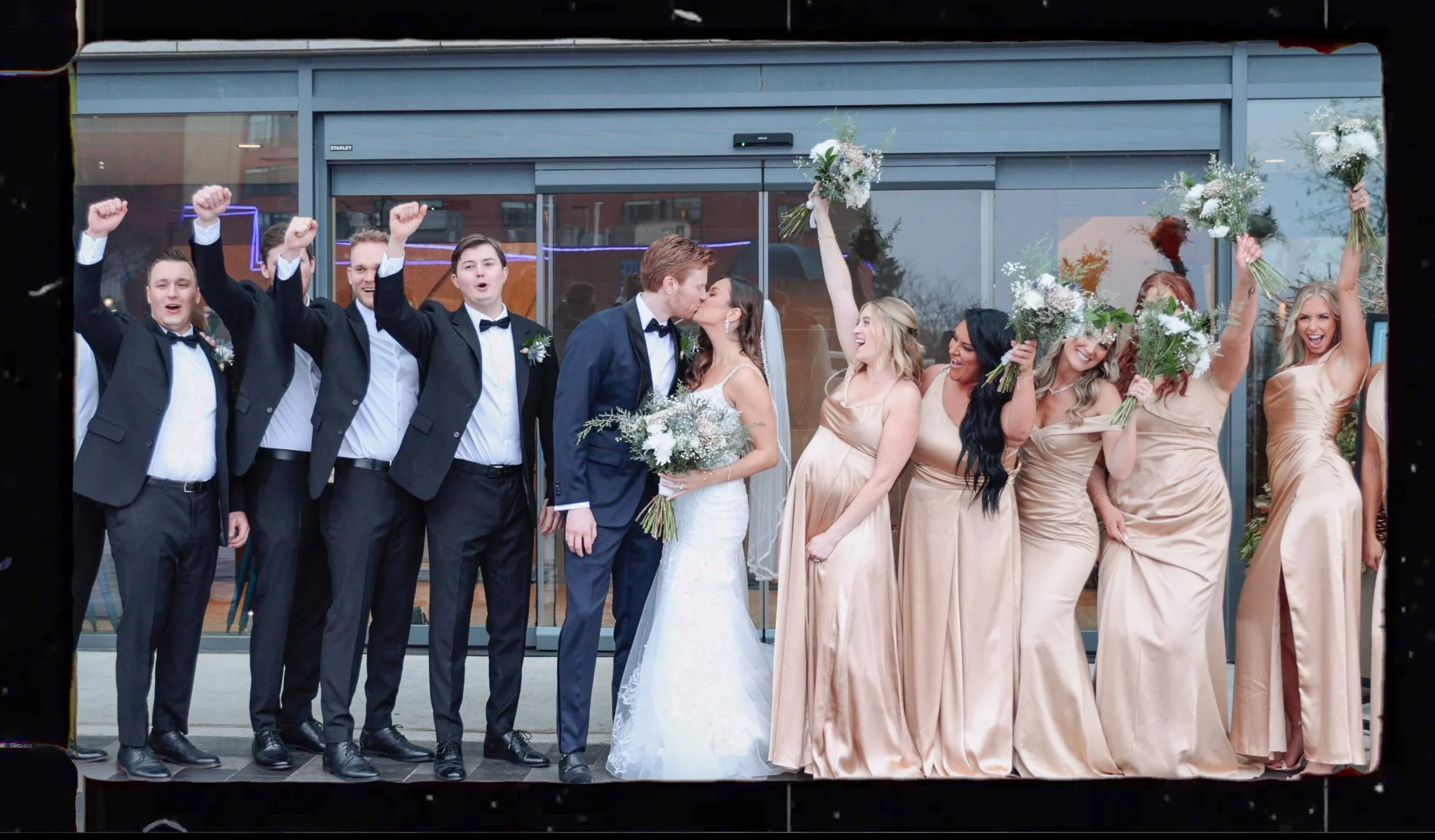 A group of wedding guests and the bride and groom celebrating outside a building. The groom and bride kiss, while the guests cheer, smile, and hold bouquets.