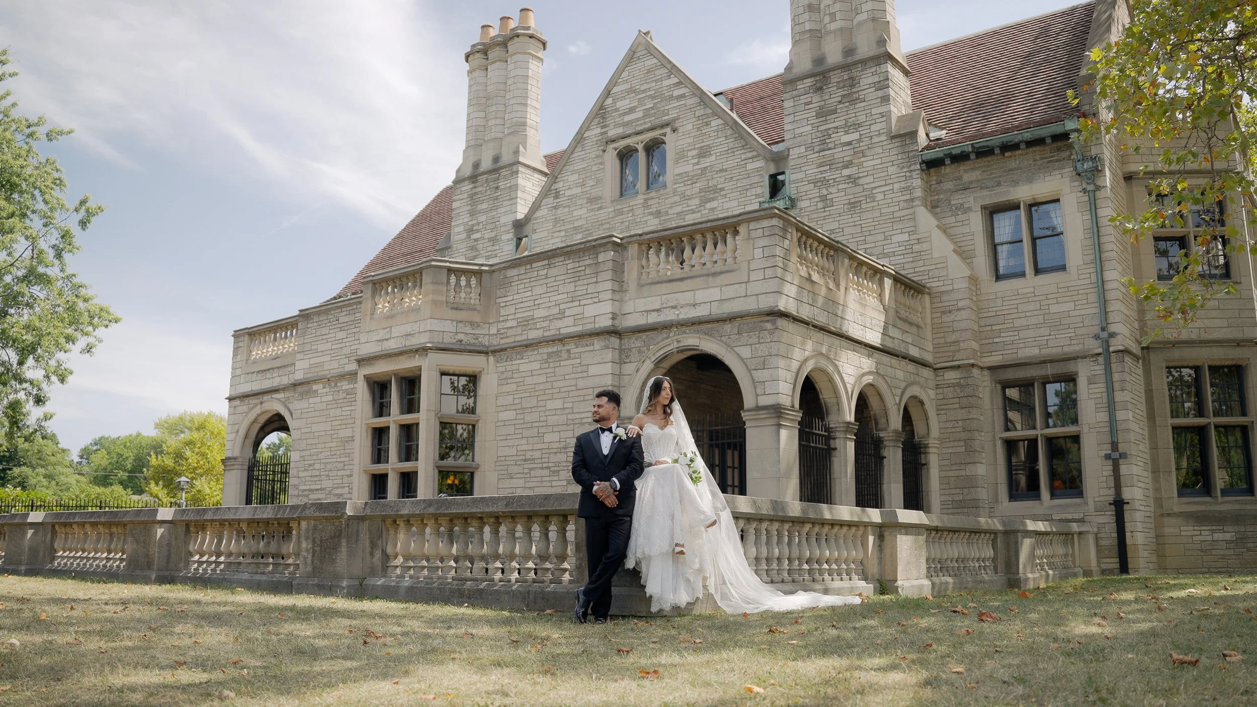A bride and groom standing in front of a large stone castle-like building with arched windows and a balcony. The groom is dressed in a black tuxedo, and the bride is wearing a white wedding gown with a long train and holding a bouquet.