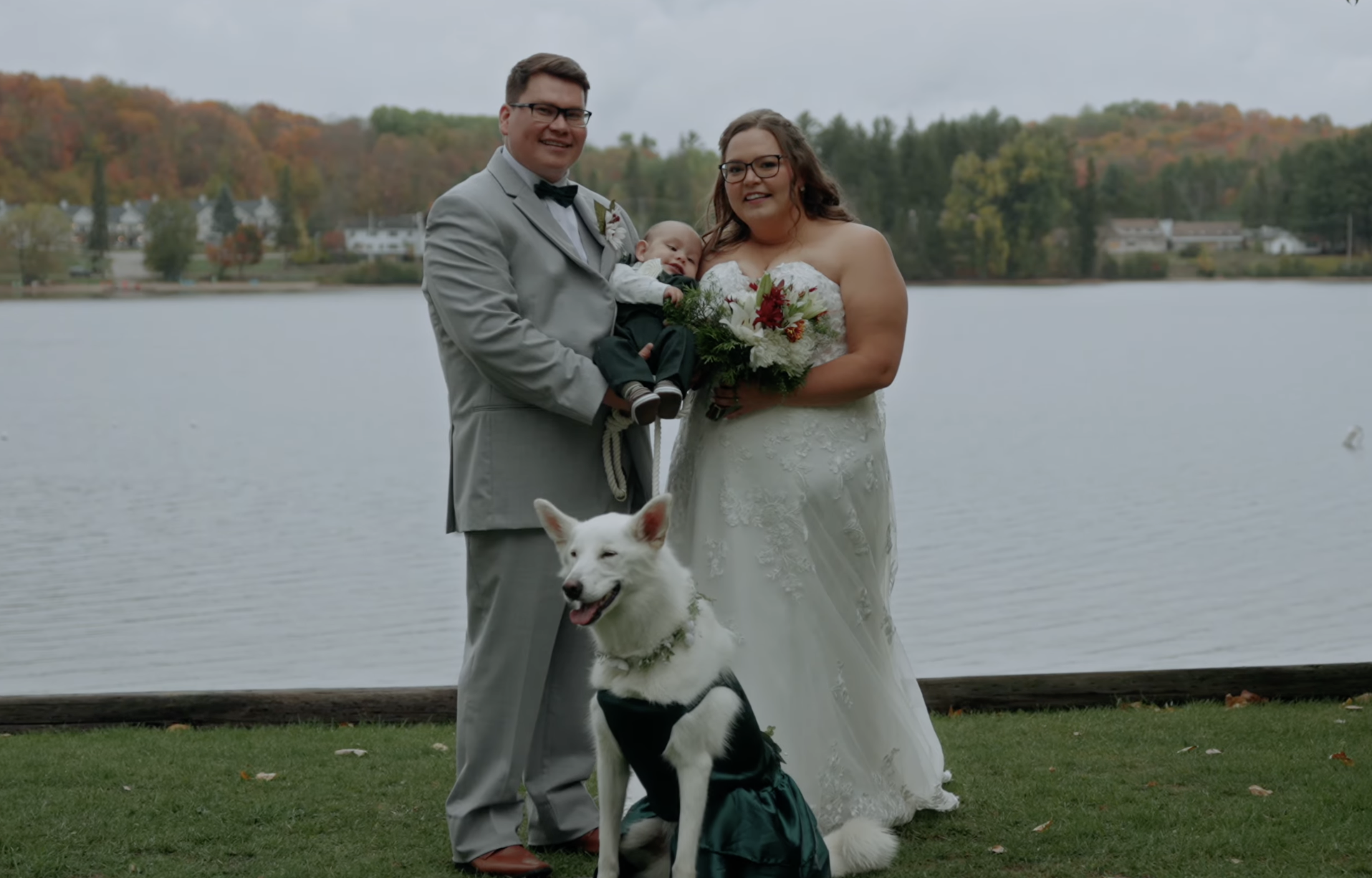 A newlywed couple standing outdoors near a lake with a dog, during autumn. The groom is in a gray suit with a black bow tie, and the bride is in a white wedding dress holding a bouquet of flowers. The groom is holding a baby dressed in a suit, and a white dog with black markings wearing a green bandana is sitting in front of them.