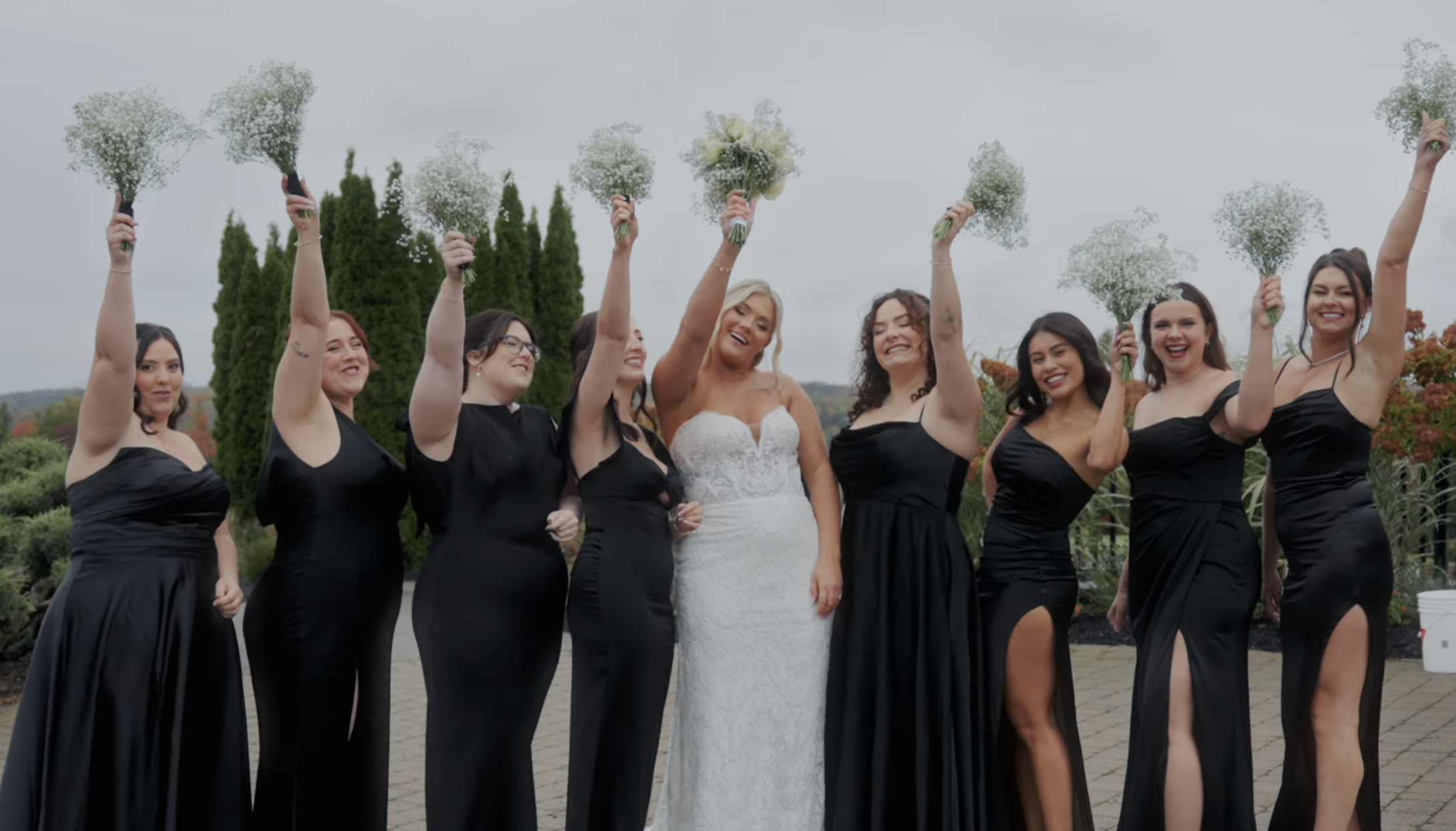 A bride in a white wedding dress surrounded by six women in black bridesmaid dresses, all smiling and holding bouquets of baby's breath flowers overhead.