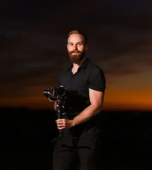 A man with a beard holding a camera stabilizer during sunset with a dark sky in the background.