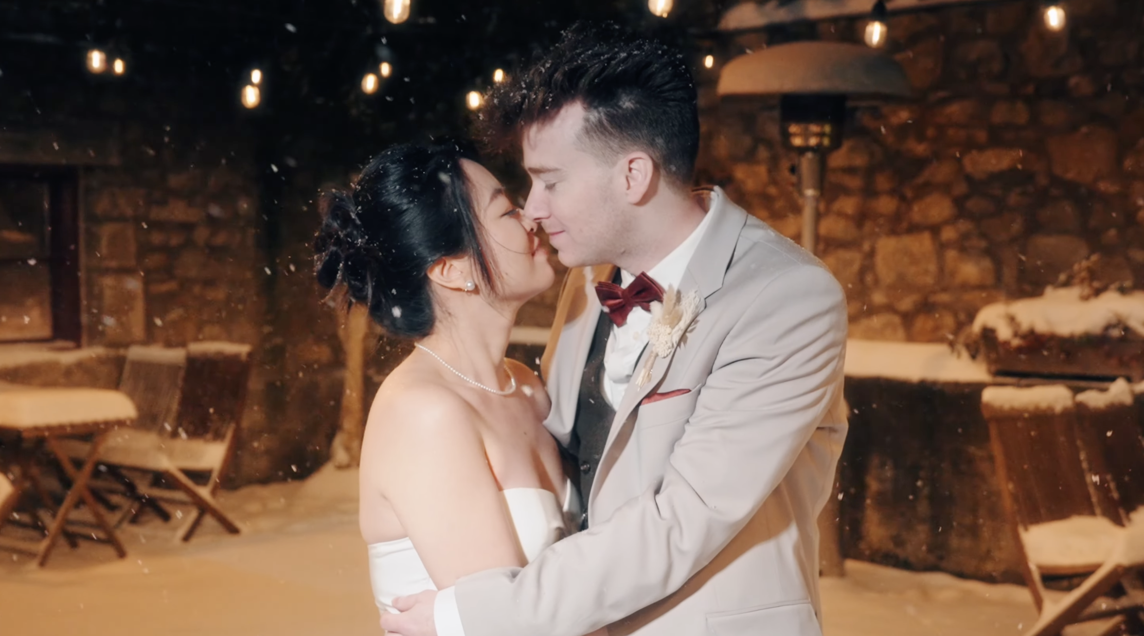 A newlywed couple dancing in the snow at night, surrounded by string lights and outdoor heaters, with snow-covered chairs in the background.