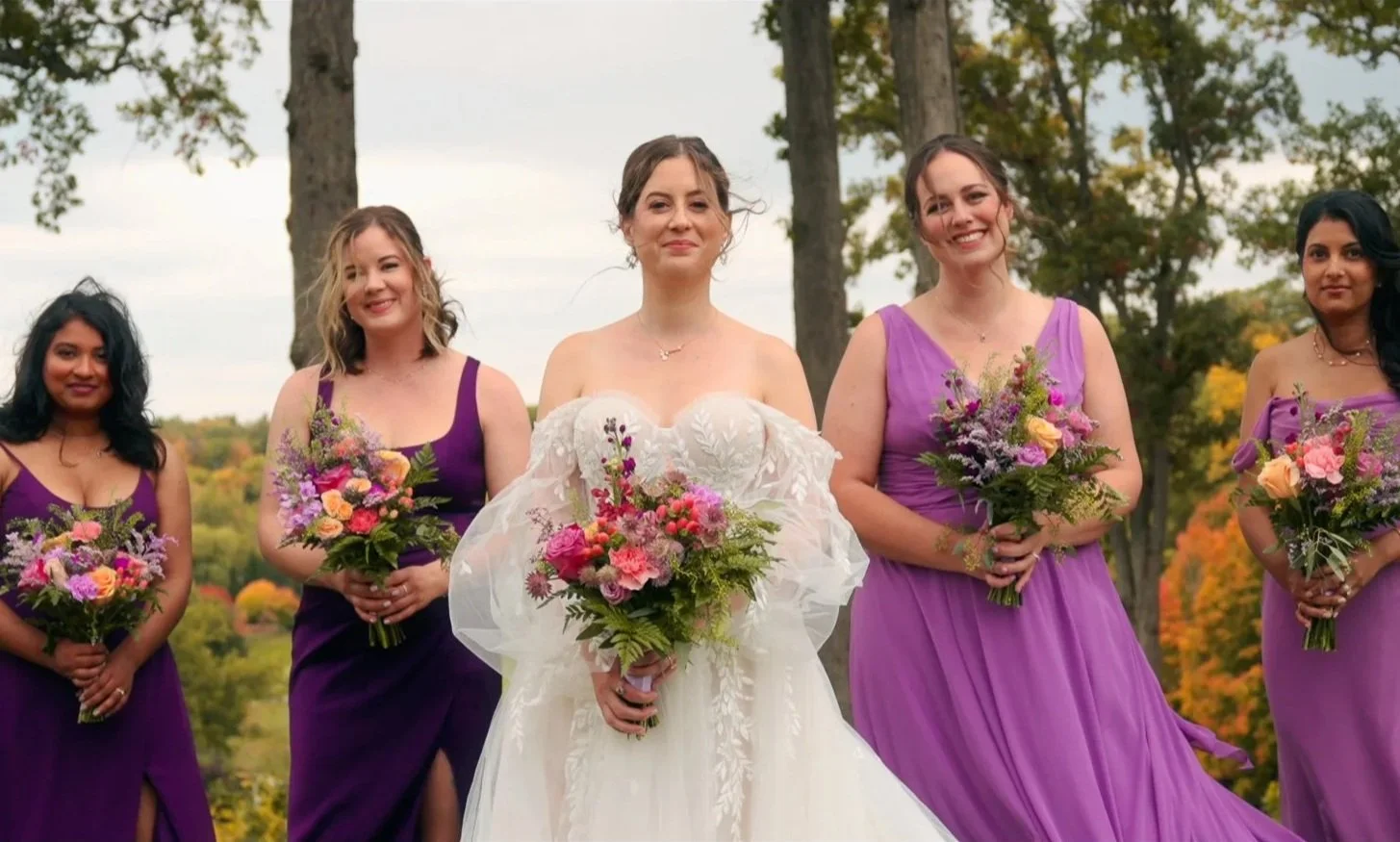 A bride and four bridesmaids standing outdoors, holding bouquets of flowers, with trees and fall foliage in the background.
