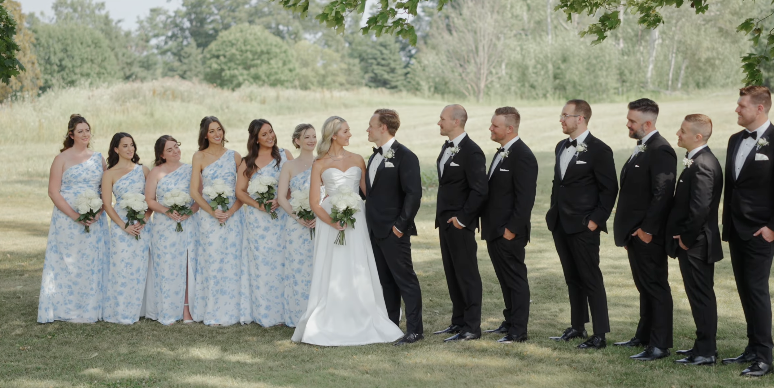 A wedding party standing outdoors on a grassy field with trees in the background, consisting of bride, groom, bridesmaids, and groomsmen facing each other with the bride and groom in the center.