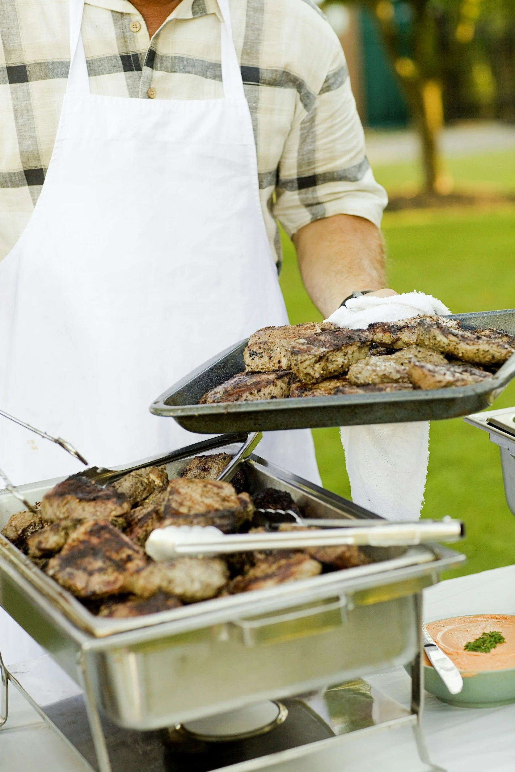 A man in a plaid shirt and white apron serving grilled ribs and meat at an outdoor barbecue.