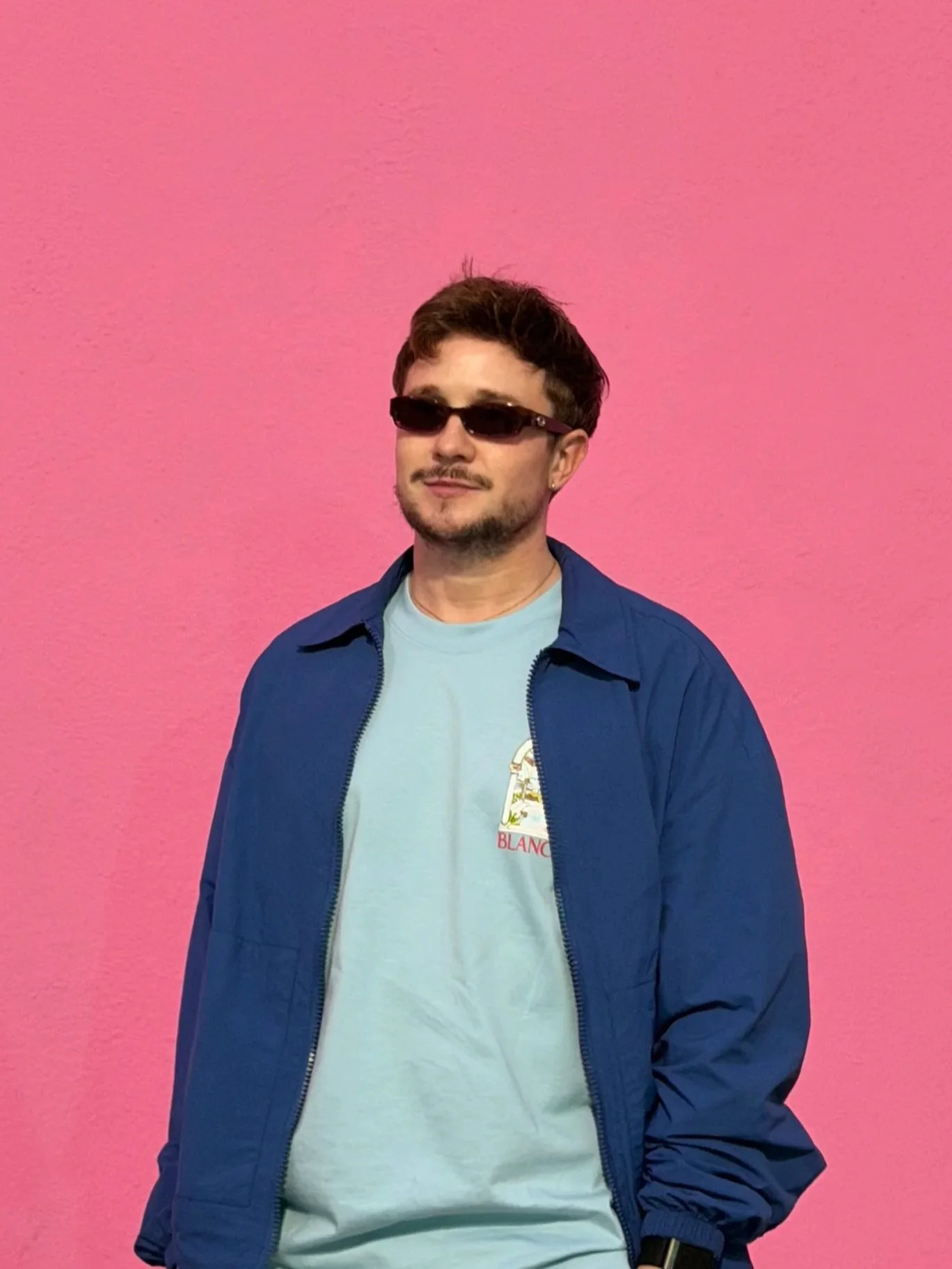 A man with short dark hair, wearing black sunglasses, a light blue t-shirt with a small logo, and a blue jacket, stands in front of a pink wall.