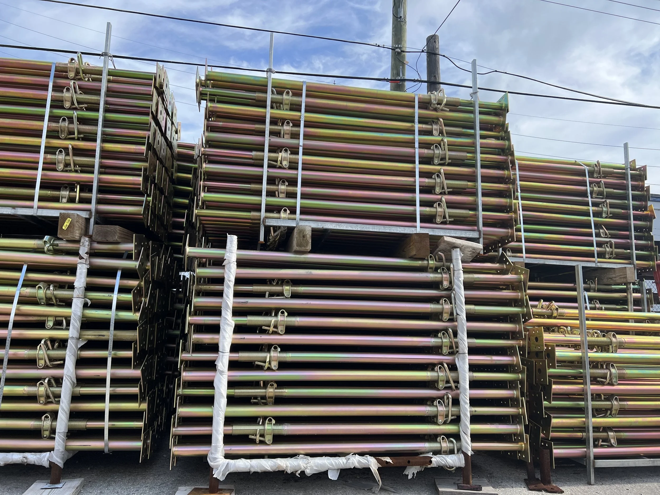 Stacked scaffolding poles and support structures stored outdoors with a cloudy sky in the background.