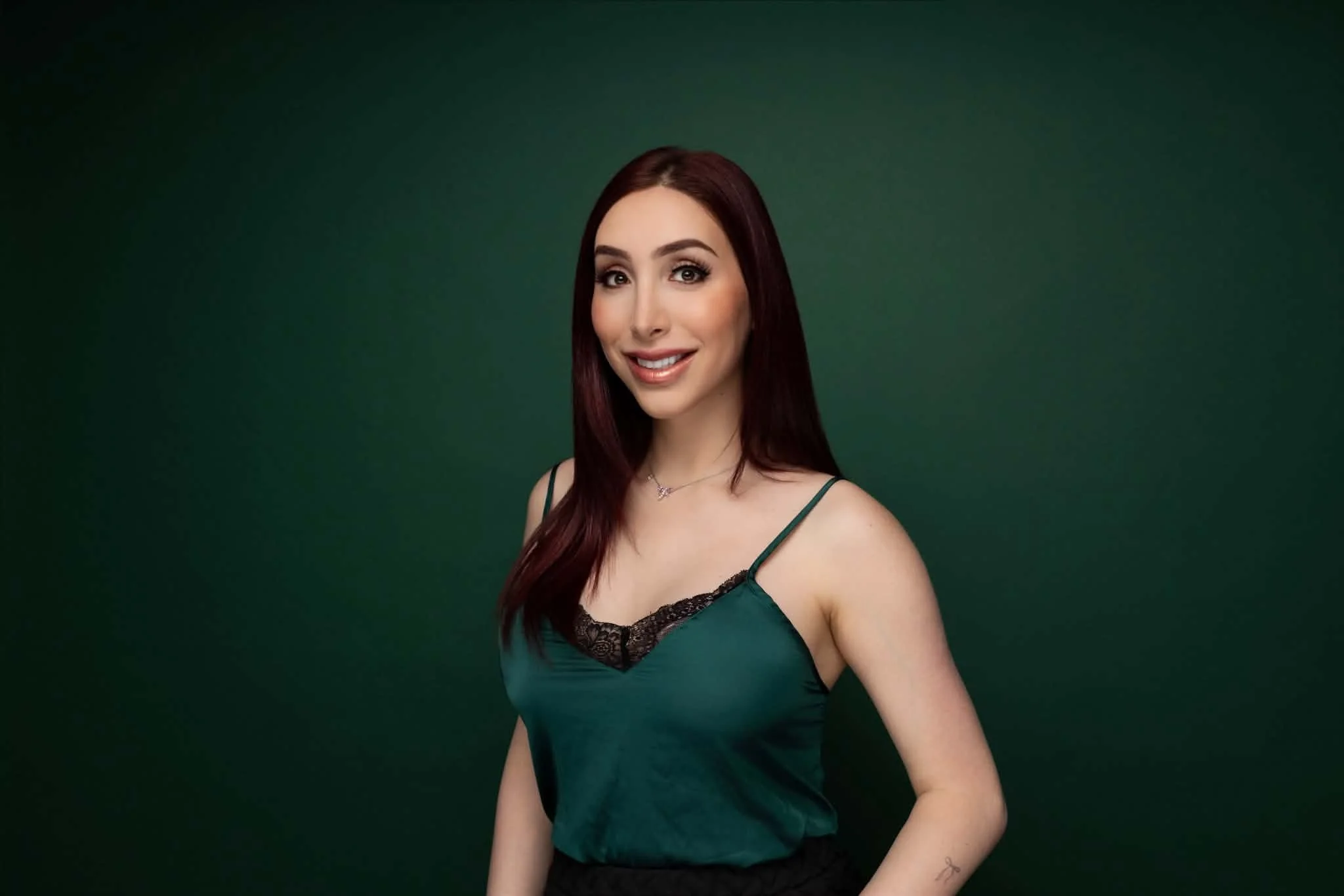 An Iranian trans woman with long, straight, red hair and soft makeup. She is wearing a green lace camisole and a black skirt.