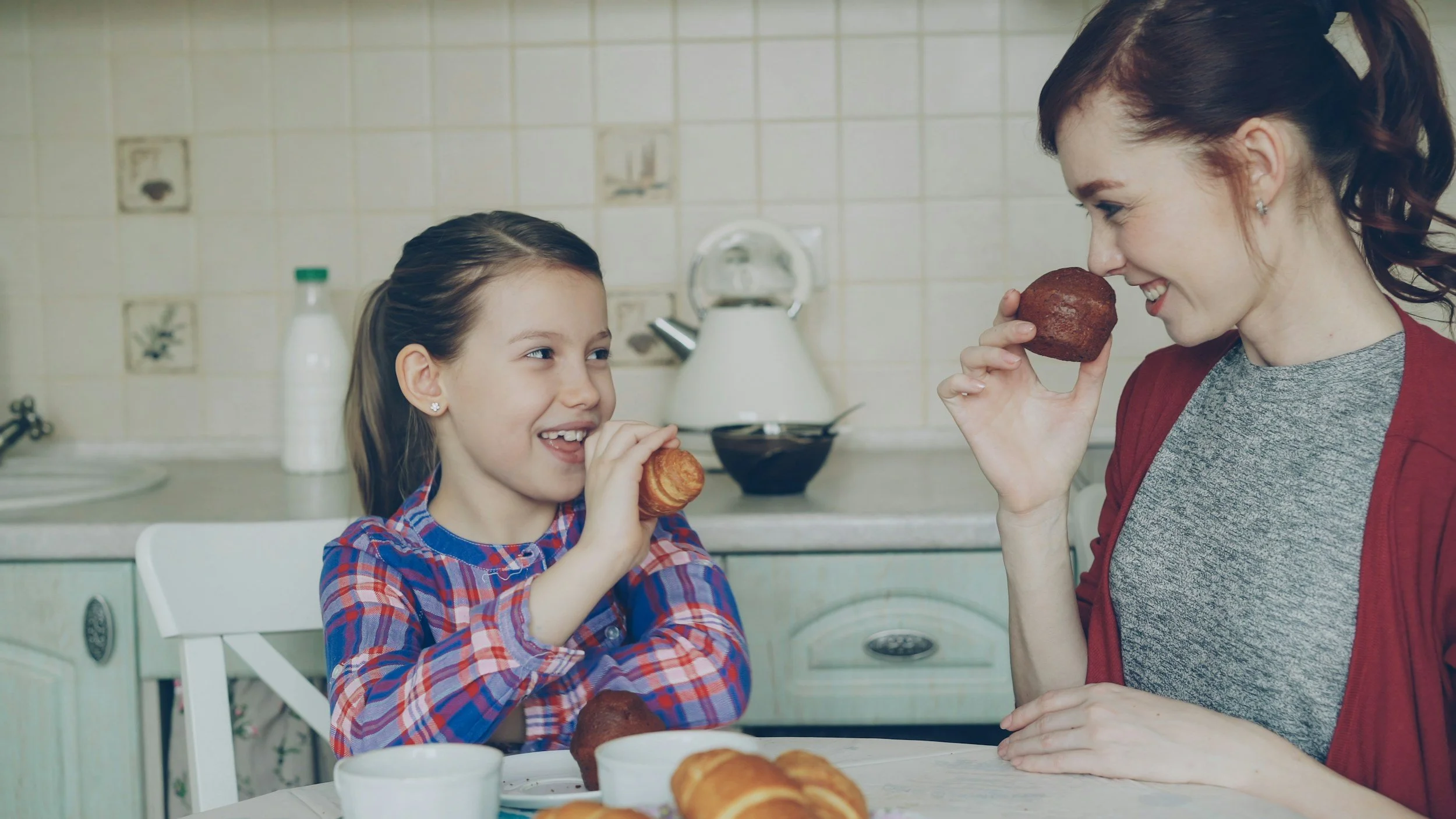 A woman and a young girl are enjoying baked goods at a kitchen table. The girl is wearing a plaid shirt and is holding a pastry near her mouth, smiling. The woman, with red hair tied back and wearing a gray shirt with a red cardigan, is holding a pastry near her nose and smiling at the girl. There are croissants and a cup on the table, with a tiled kitchen background.
