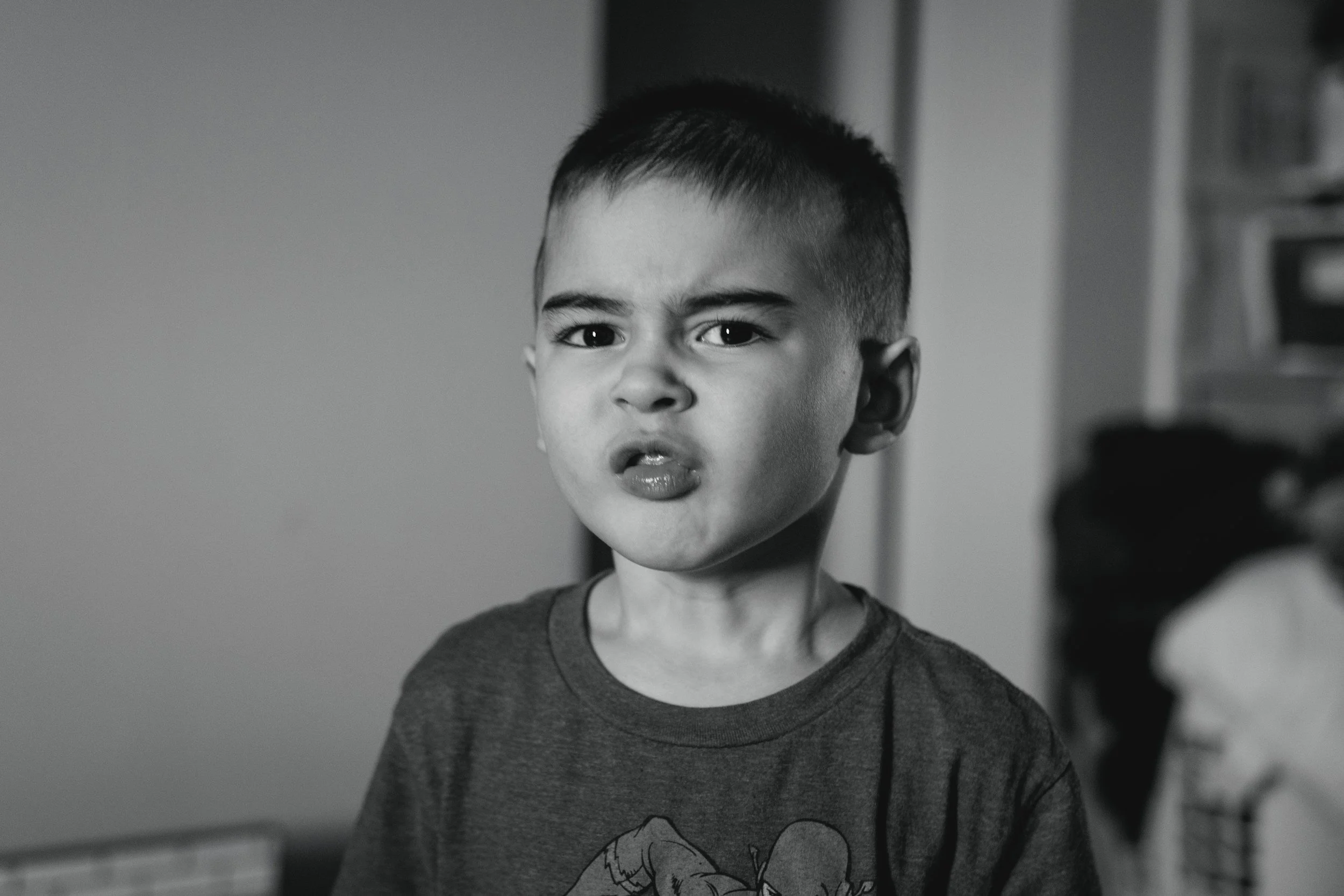 A young boy with short, dark hair and a serious expression, wearing a T-shirt with a cartoon character, looking towards the camera indoors.