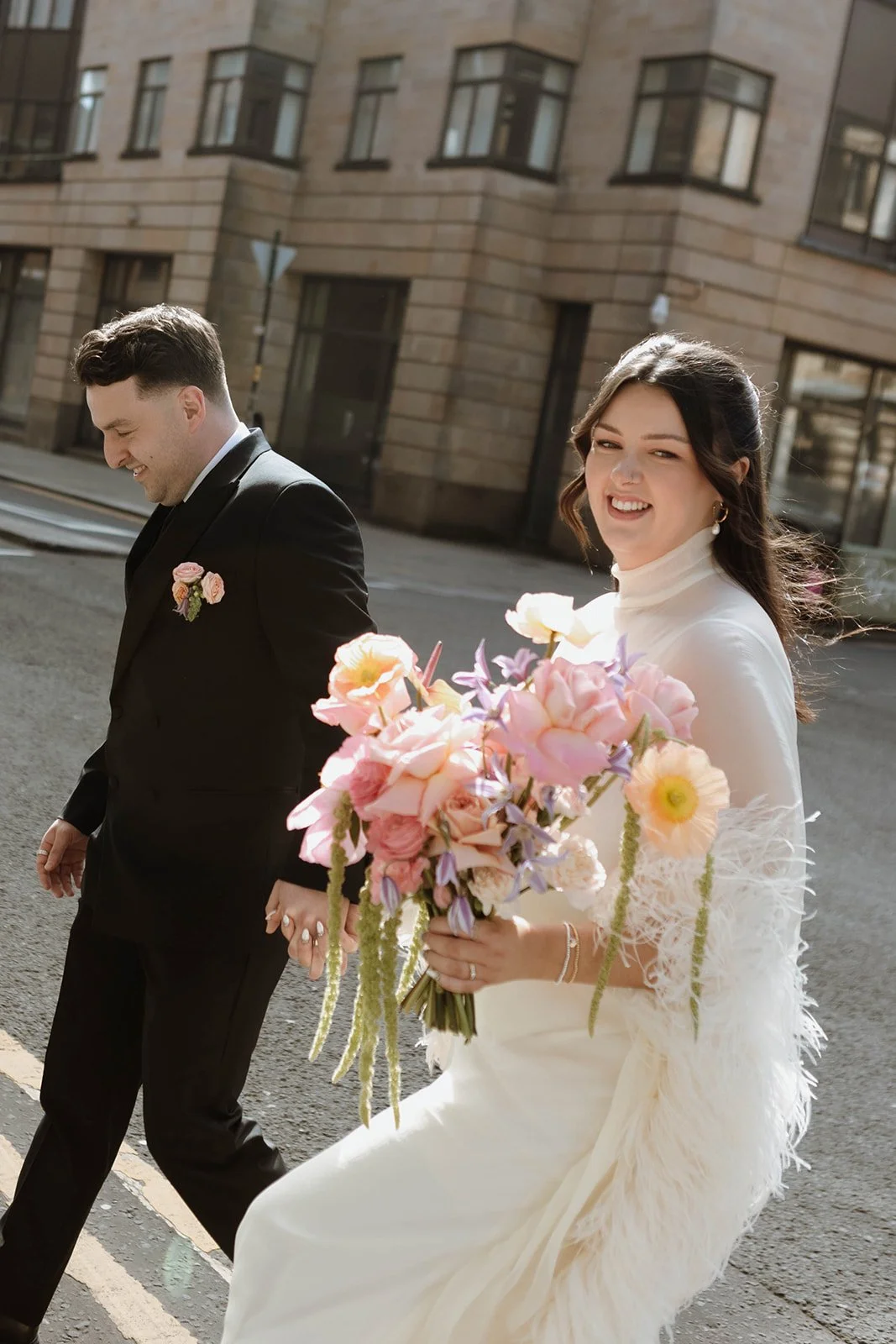 Bride in white dress holding a bouquet of pink, purple, and cream flowers, walking with groom in black suit on city street during day.