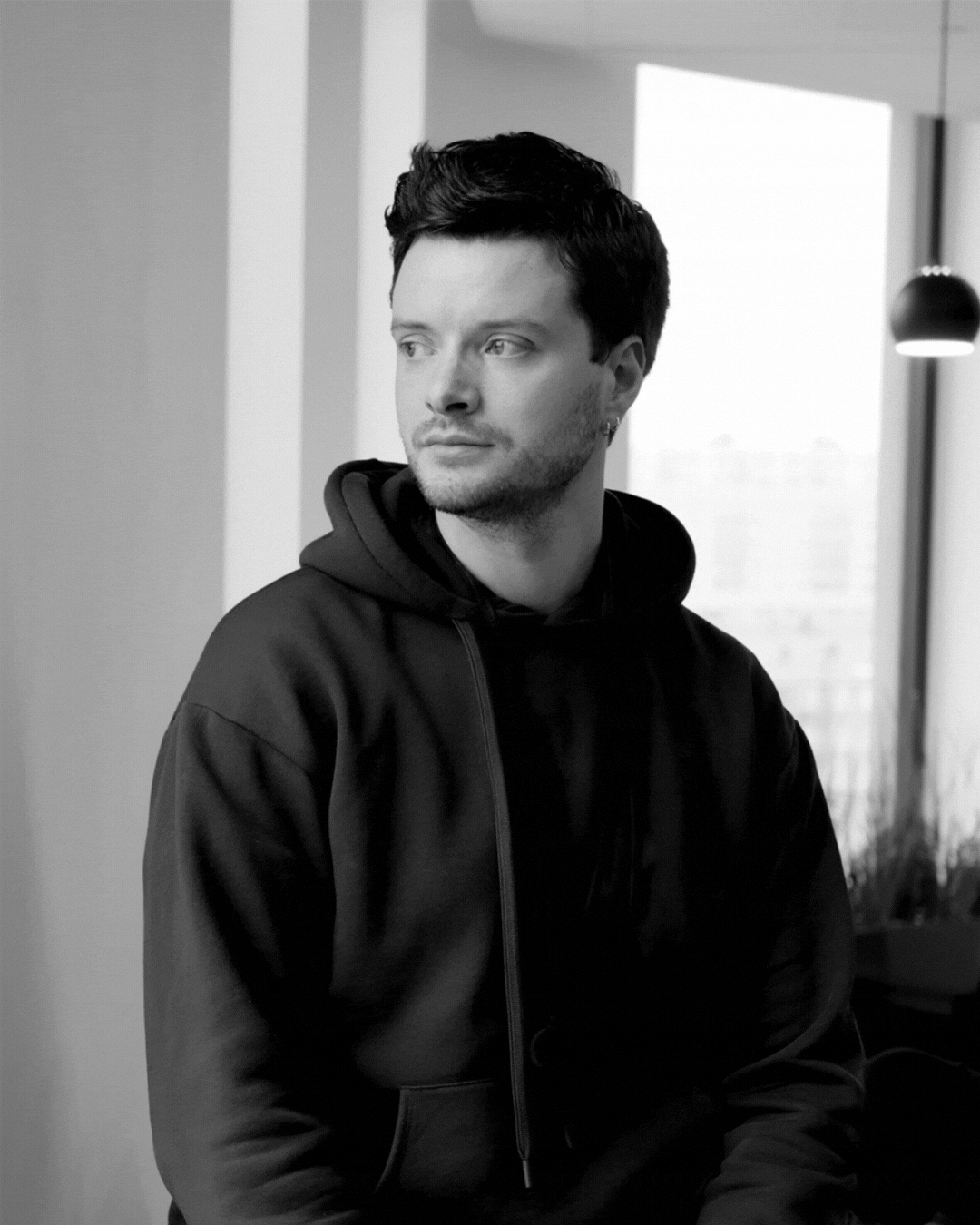 A young man with dark hair and light skin, wearing a black hoodie, looking to the side inside a modern indoor space with a window and a hanging lamp in the background.