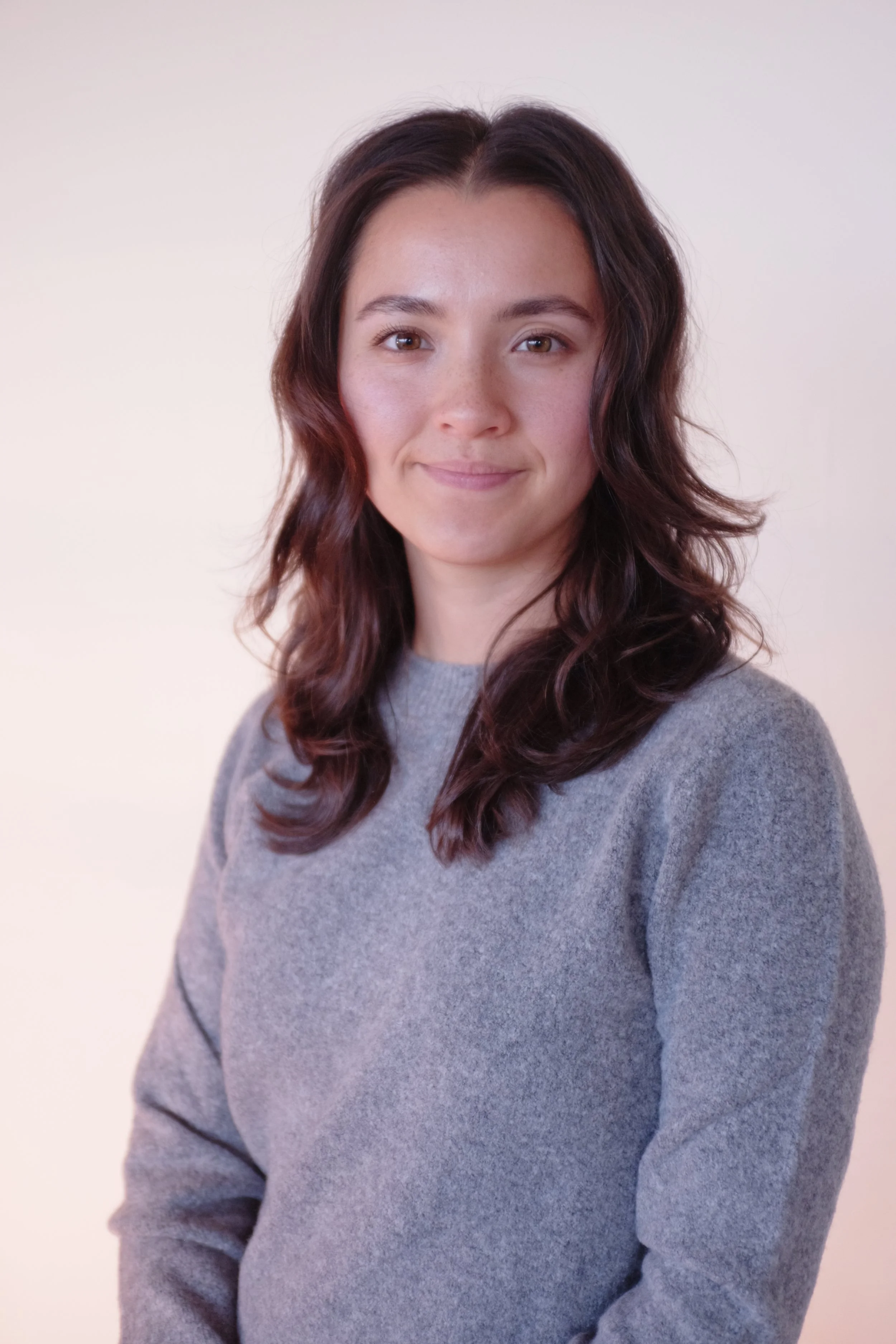 Portrait of a young woman with shoulder-length wavy dark brown hair, wearing a light gray sweater, smiling softly, against a plain beige background.