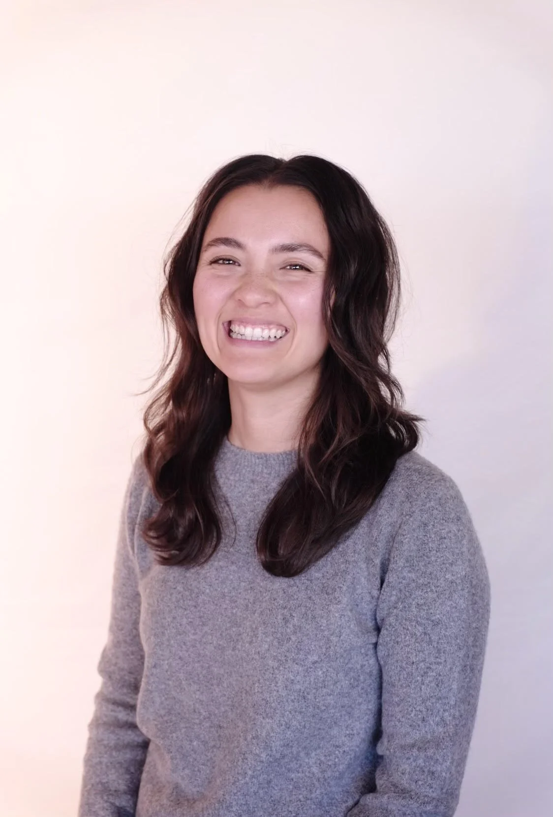 A smiling woman with dark wavy hair wearing a gray sweater, standing against a plain light background.