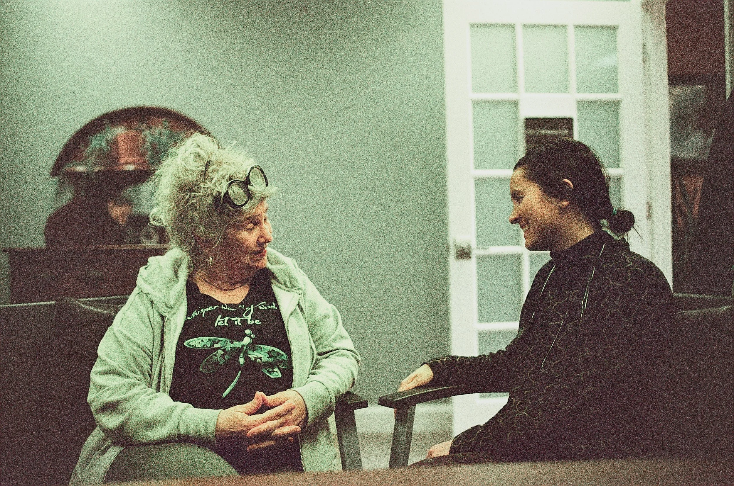 Two women smiling and talking in a living room. The older woman has curly gray hair, glasses on her head, and is wearing a black T-shirt with a design and a light green hoodie. The younger woman has dark hair tied back and is wearing a dark patterned top. They are seated facing each other with a wooden table between them, and there is a door and a mirror in the background.
