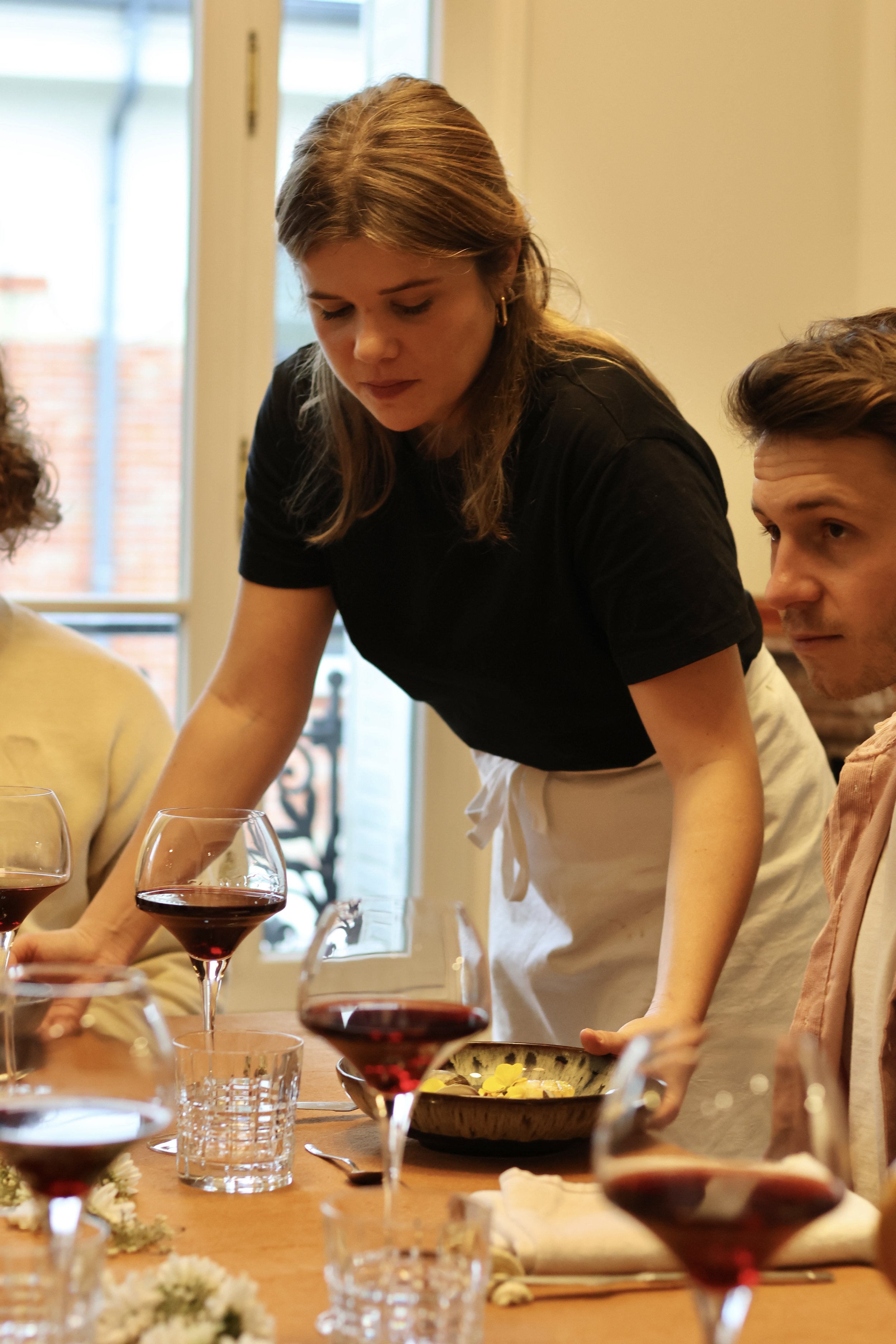Une femme serveuse sert du vin rouge à un groupe de personnes lors d'un repas dans une salle lumineuse avec une fenêtre en arrière-plan.