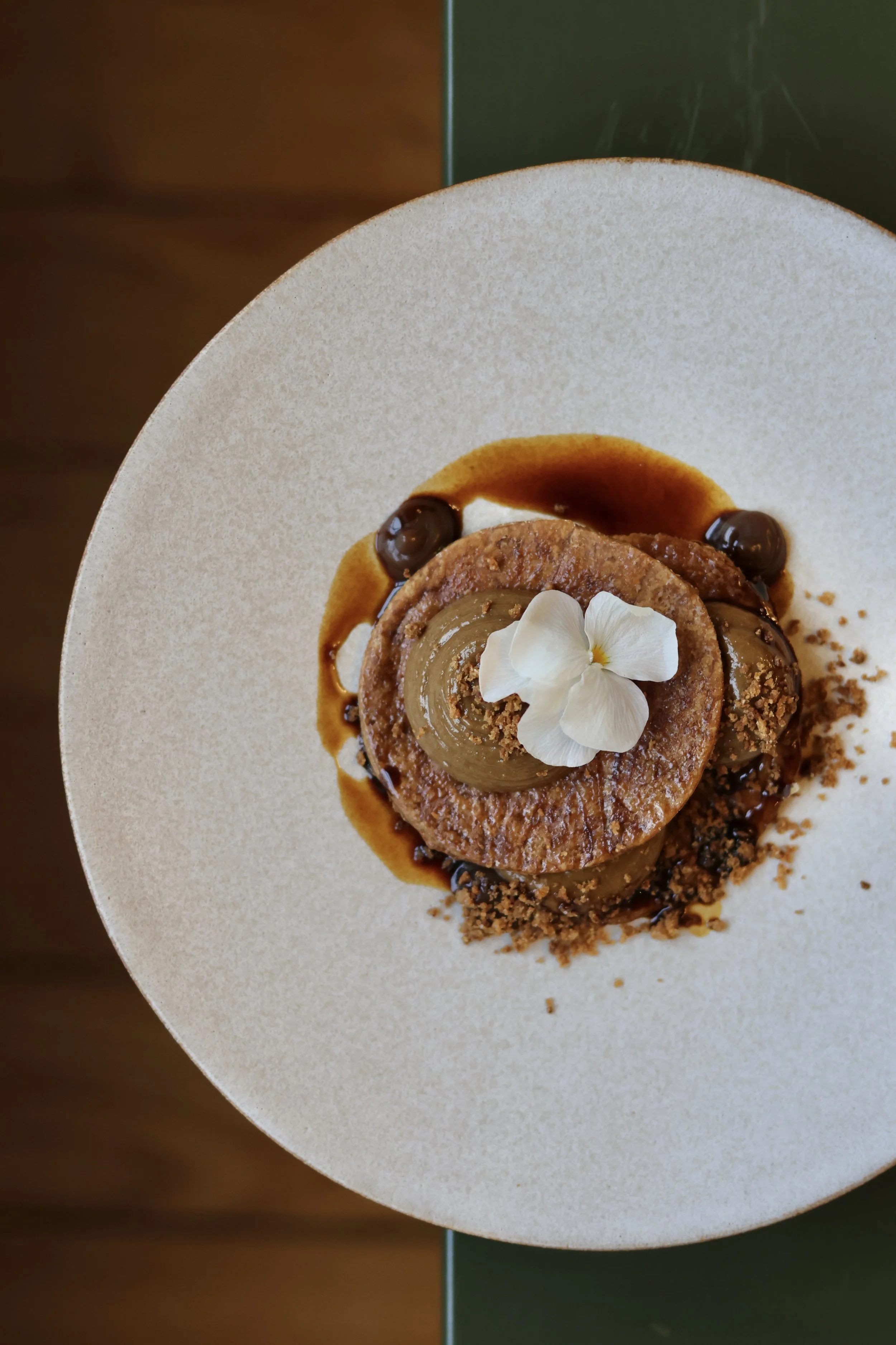 Un dessert présenté sur une assiette blanche. Il comprend un gâteau rond au centre, décoré de fleurs blanches, avec une sauce caramel et des décorations en chocolat autour.