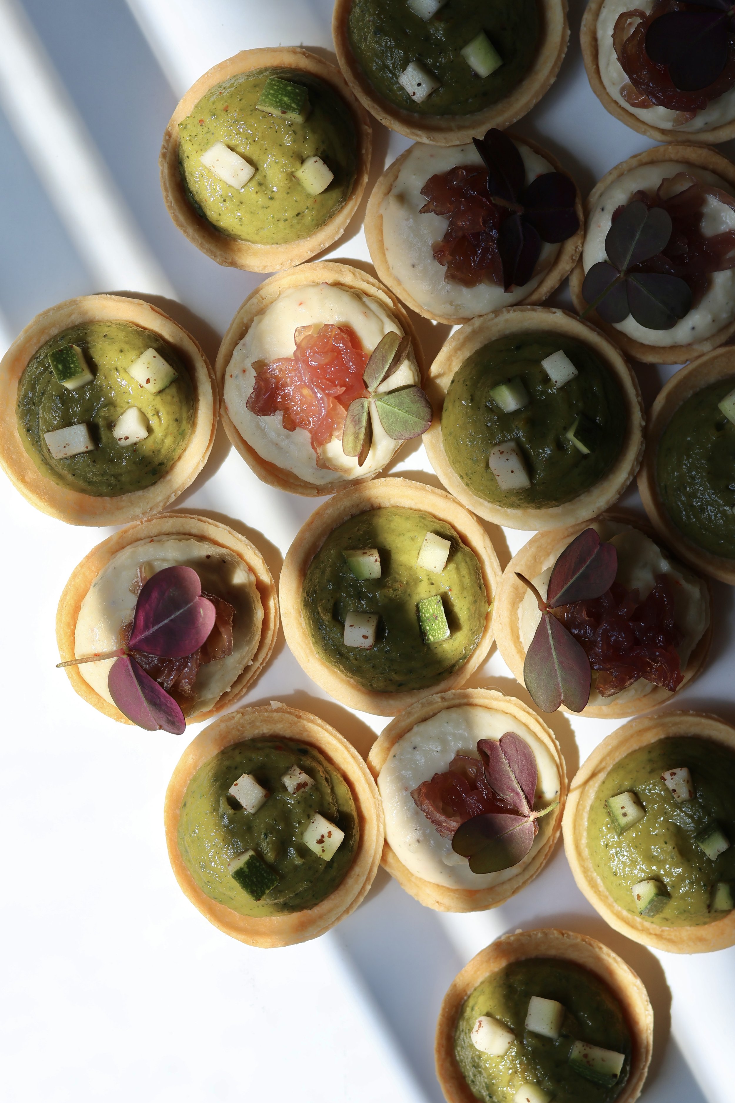 Assortiment de tartelettes apéritives avec différentes garnitures, dont du pesto, de la crème et des morceaux de légumes ou de viande, décorées de petites feuilles de basilic ou autre herbes.