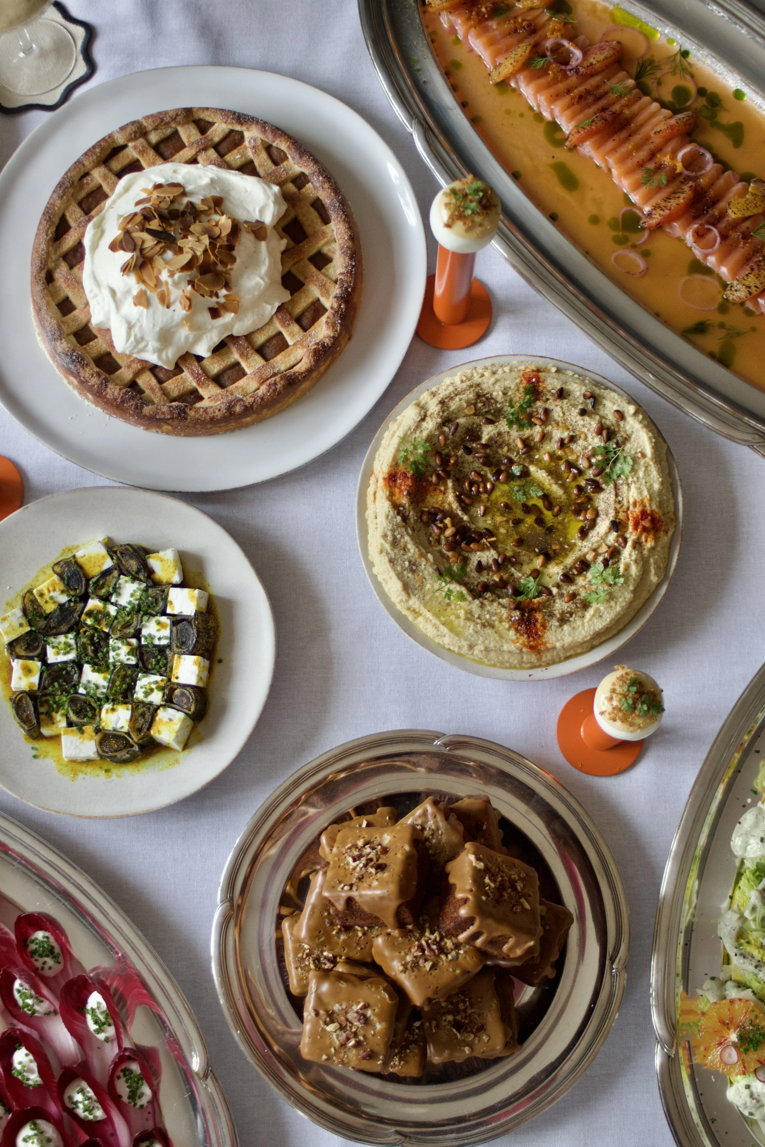 Plateau de plats méditerranéens et desserts, y compris houmous, salade de tomates, tartes, et desserts sucrés en vitrine, présentation élégante sur nappe blanche.