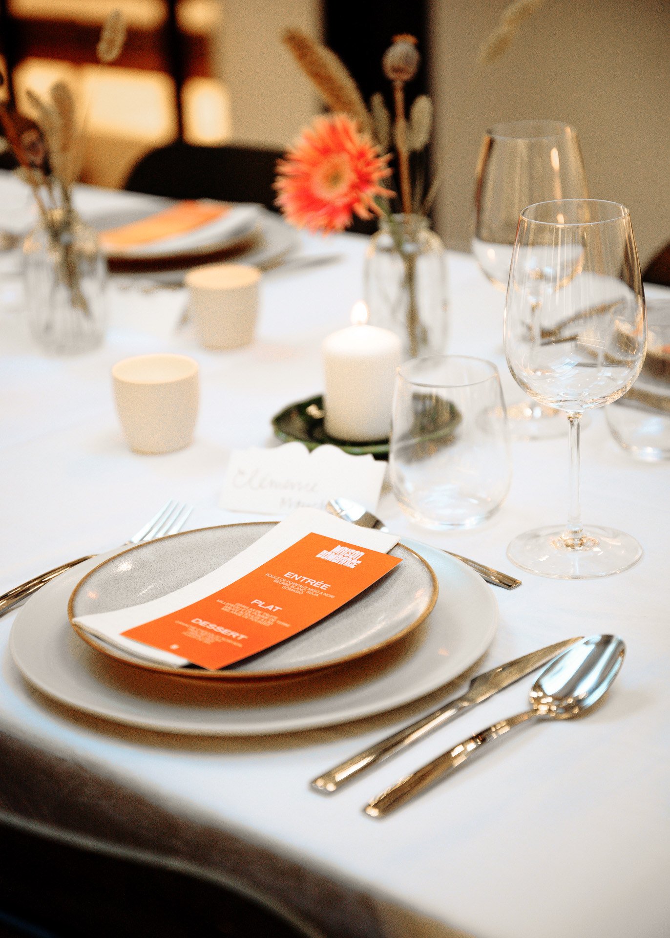 Une table élégante prête pour un repas, avec des assiettes, des verres, des couverts, une bougie et un menu orange, décorée avec des fleurs.
