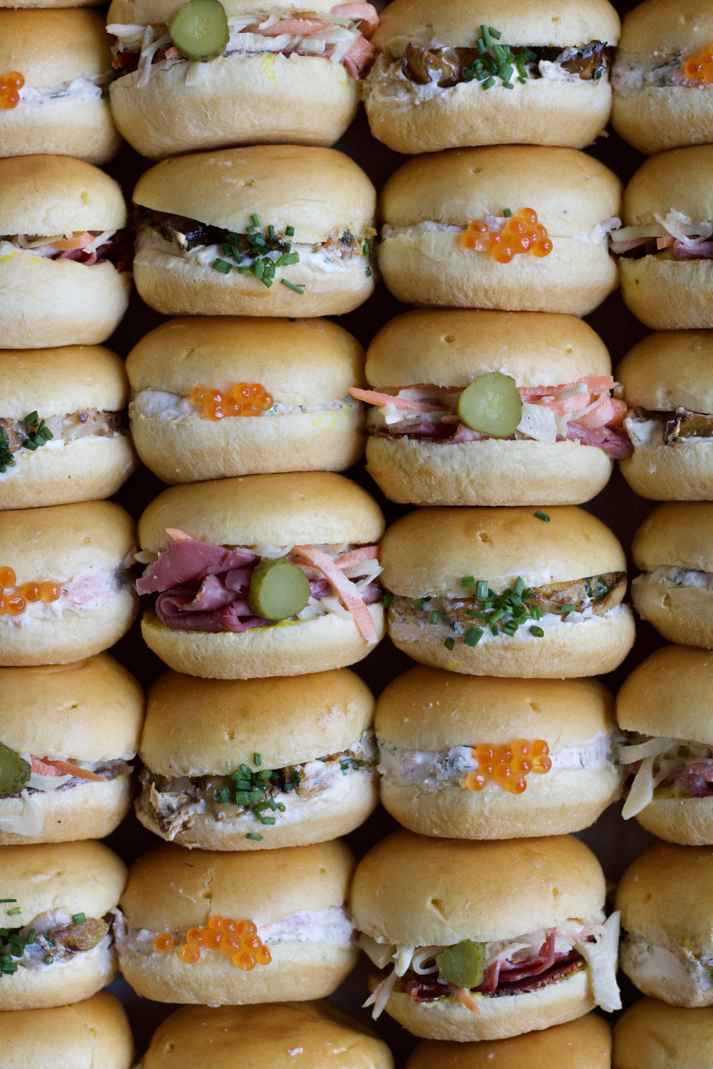 Une rangée de mini sandwiches ou bouchées apéritives, garnis de diverses garnitures comme du saumon, du jambon, des pickles, des caviar d'algues, et des herbes, servis sur des petits pains ronds.